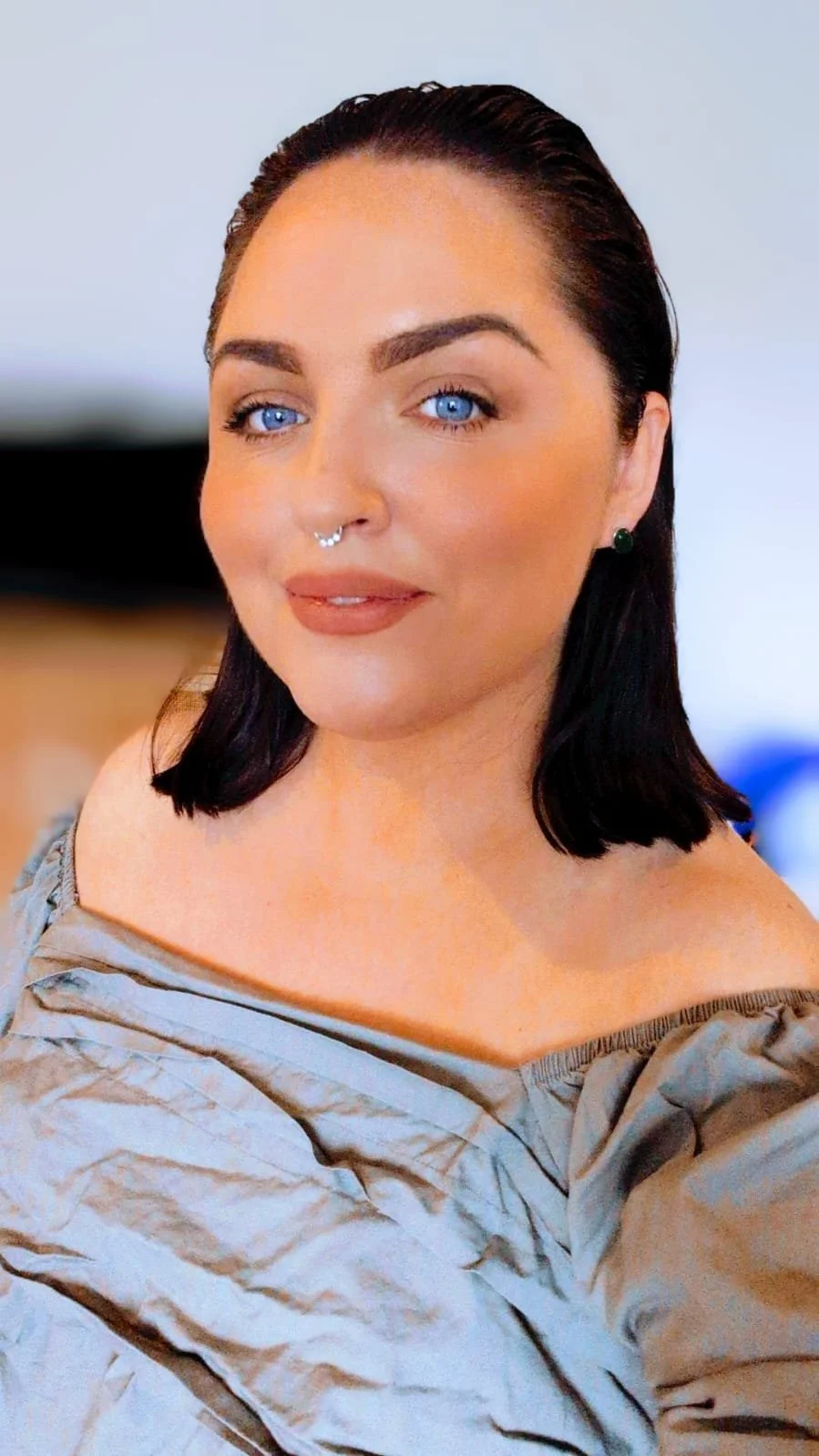 Close-up portrait of a woman with blue eyes, dark brown hair, and a septum piercing, wearing a light-colored off-shoulder top.