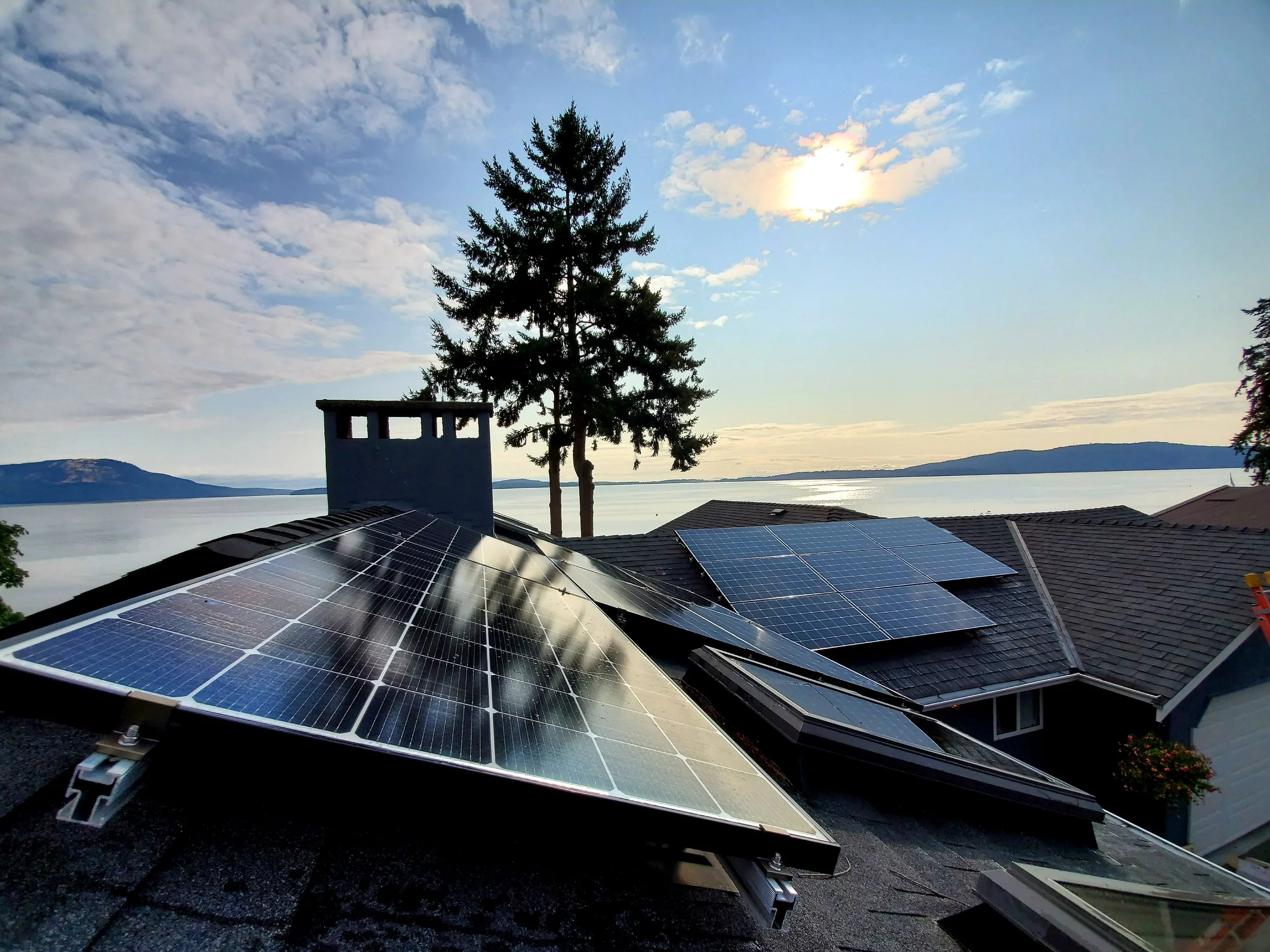 Residential rooftops with solar panels, a pine tree, and a chimney, overlooking a lake with mountains in the distance at sunset or sunrise.
