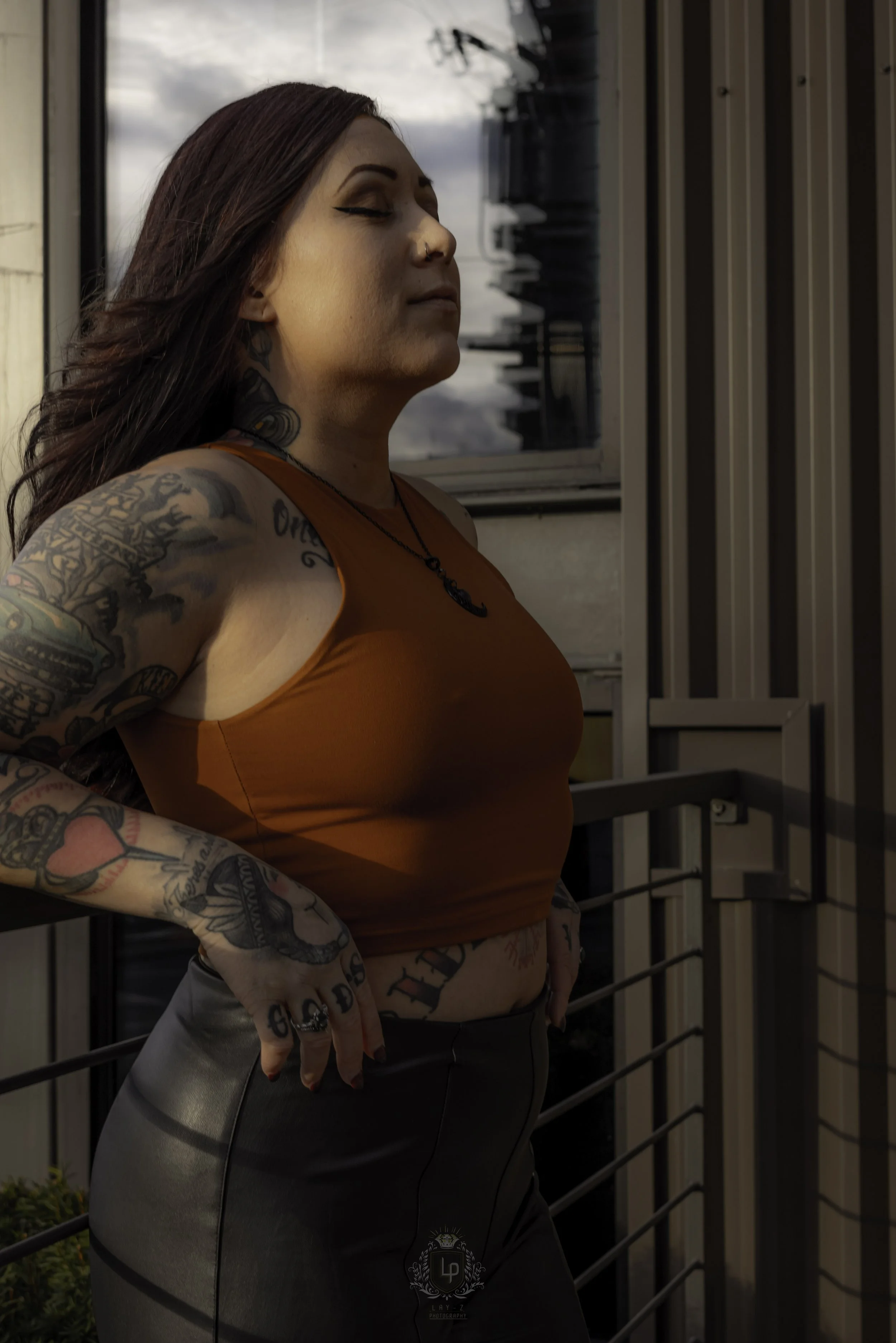Woman with tattoos standing outside, eyes closed, wearing an orange sleeveless top and black pants, with her hands resting on her hips.