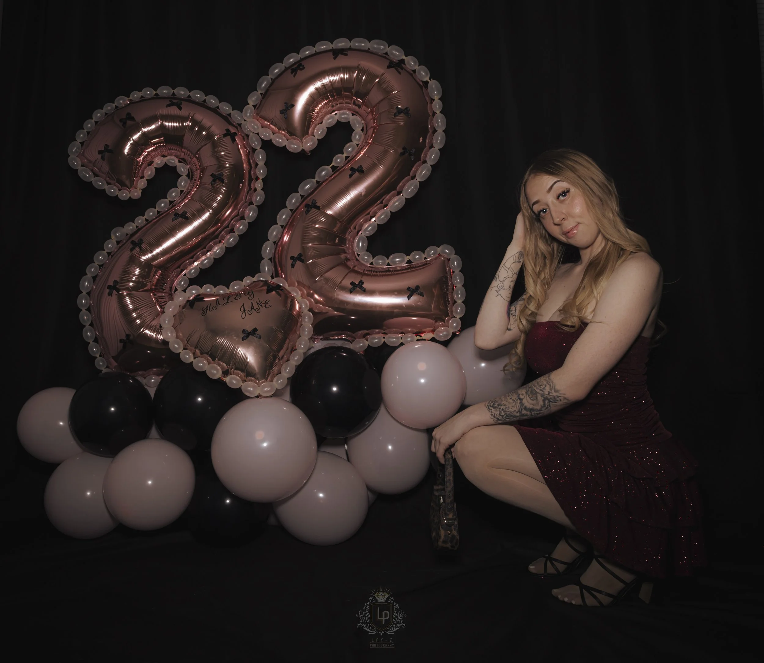 A young woman with long blonde hair, wearing a sparkly red dress and black high-heeled shoes, is sitting on the floor near a display of large pink and black balloons. The balloons include two large metallic pink number-shaped balloons forming a '22',