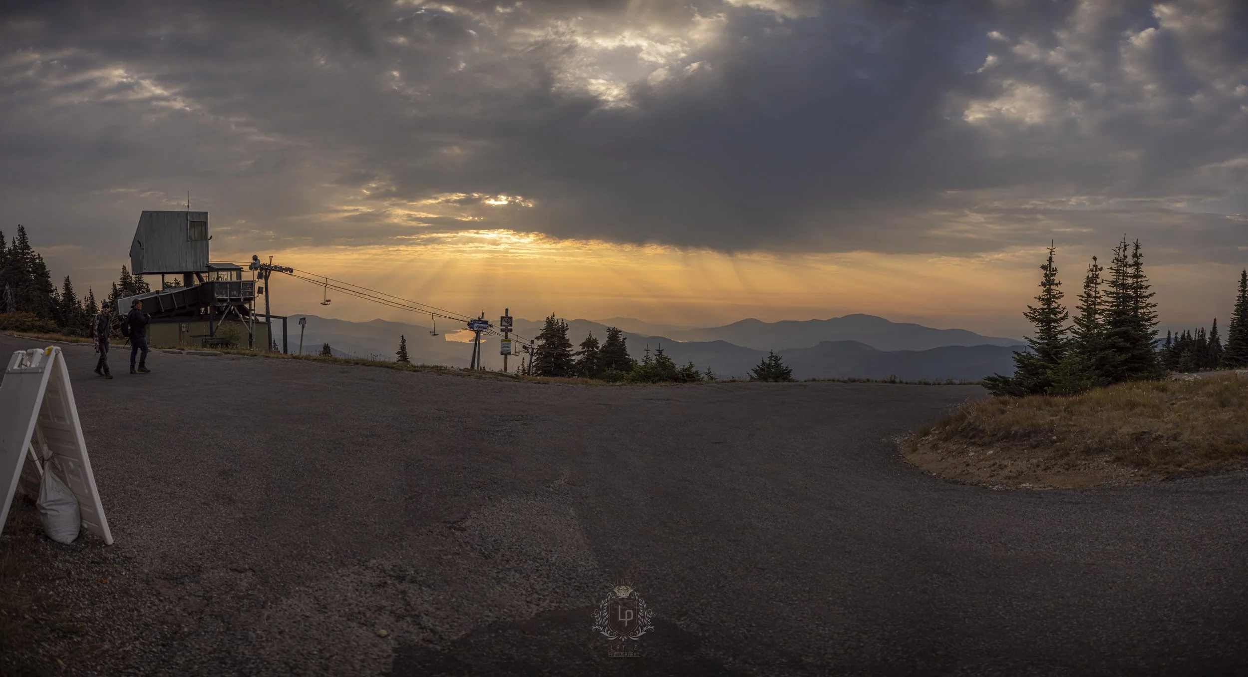 Mountain landscape during sunset with a cloudy sky, pine trees, and a ski lift station with a few people nearby.