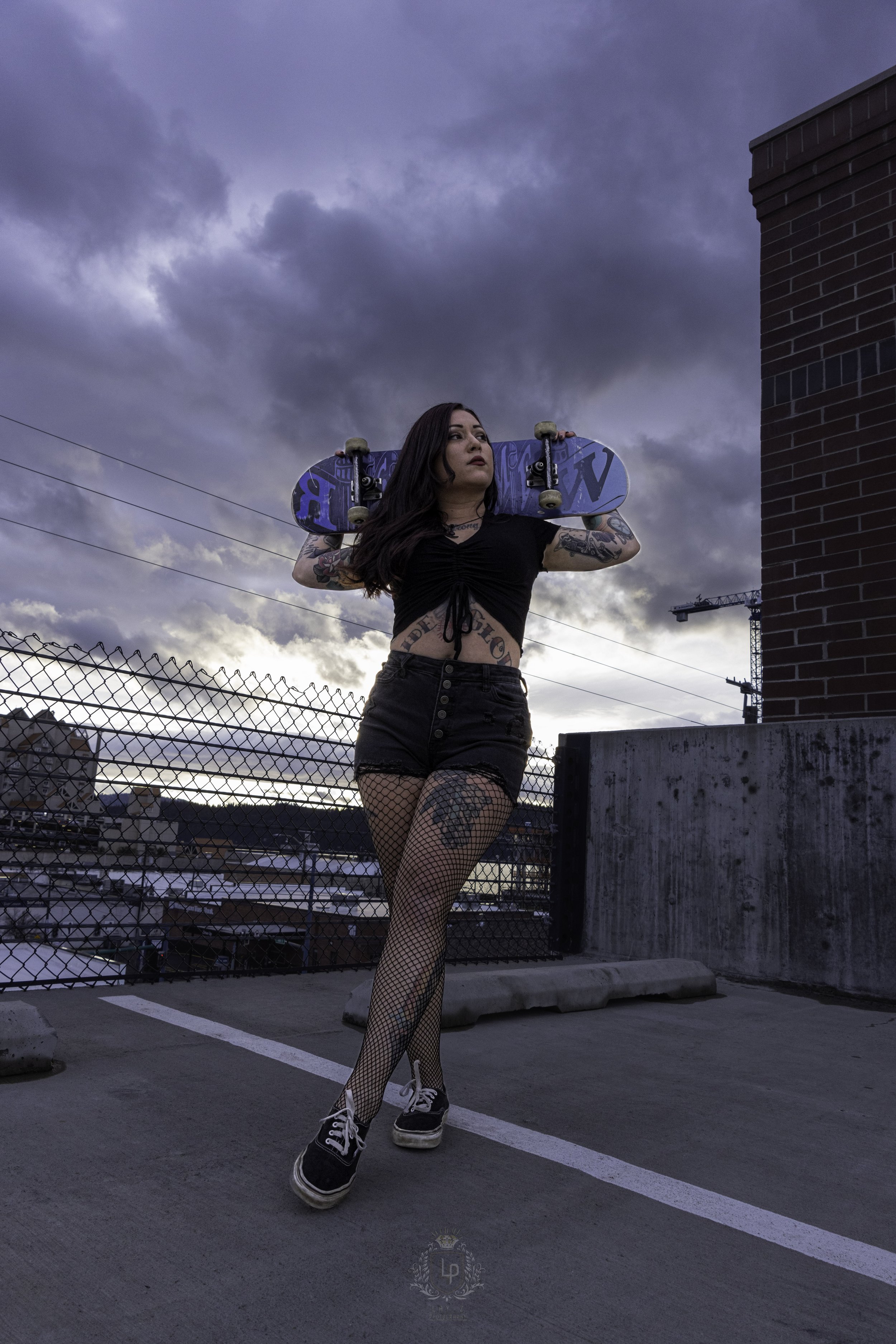 A woman stands on a parking garage rooftop holding a skateboard on her shoulders, wearing a black crop top, black shorts, fishnet stockings, and sneakers, with a cloudy sky behind her.