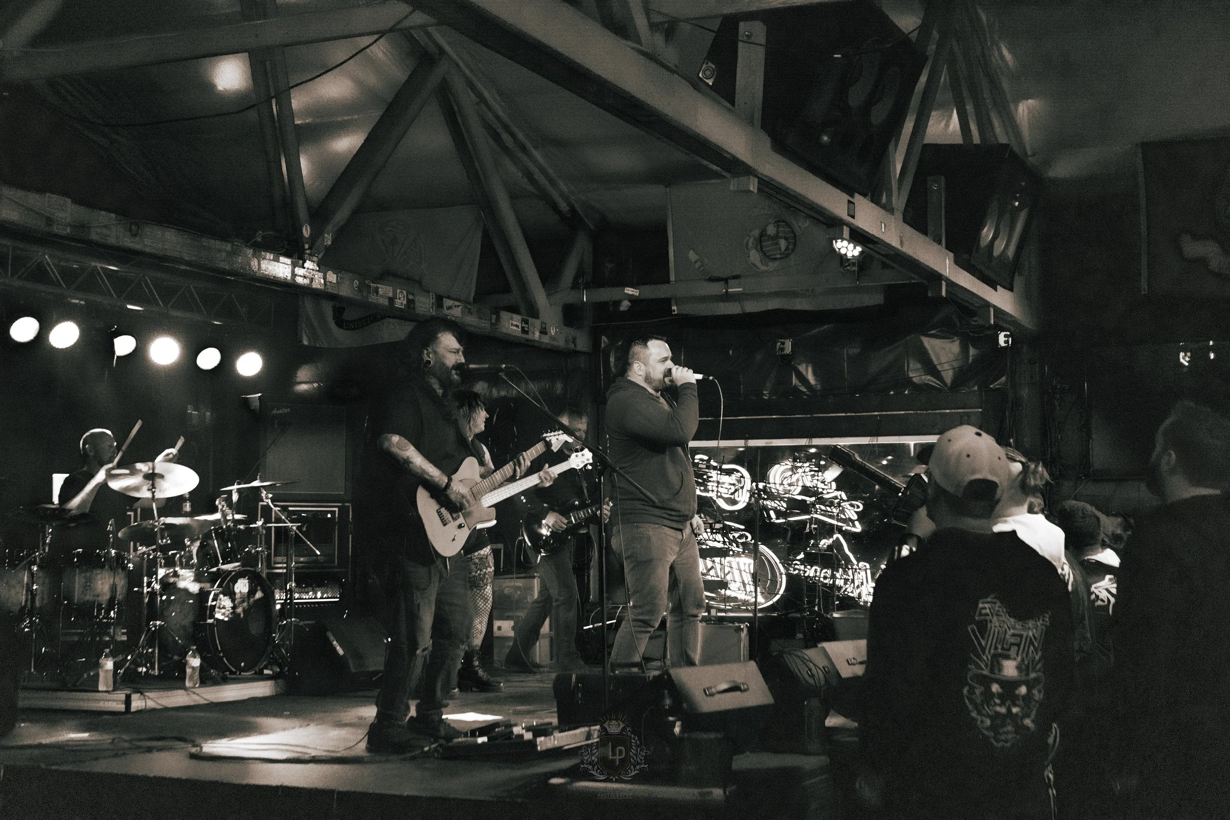 Black and white photo of a live band performing on stage at a venue, with audience members watching.