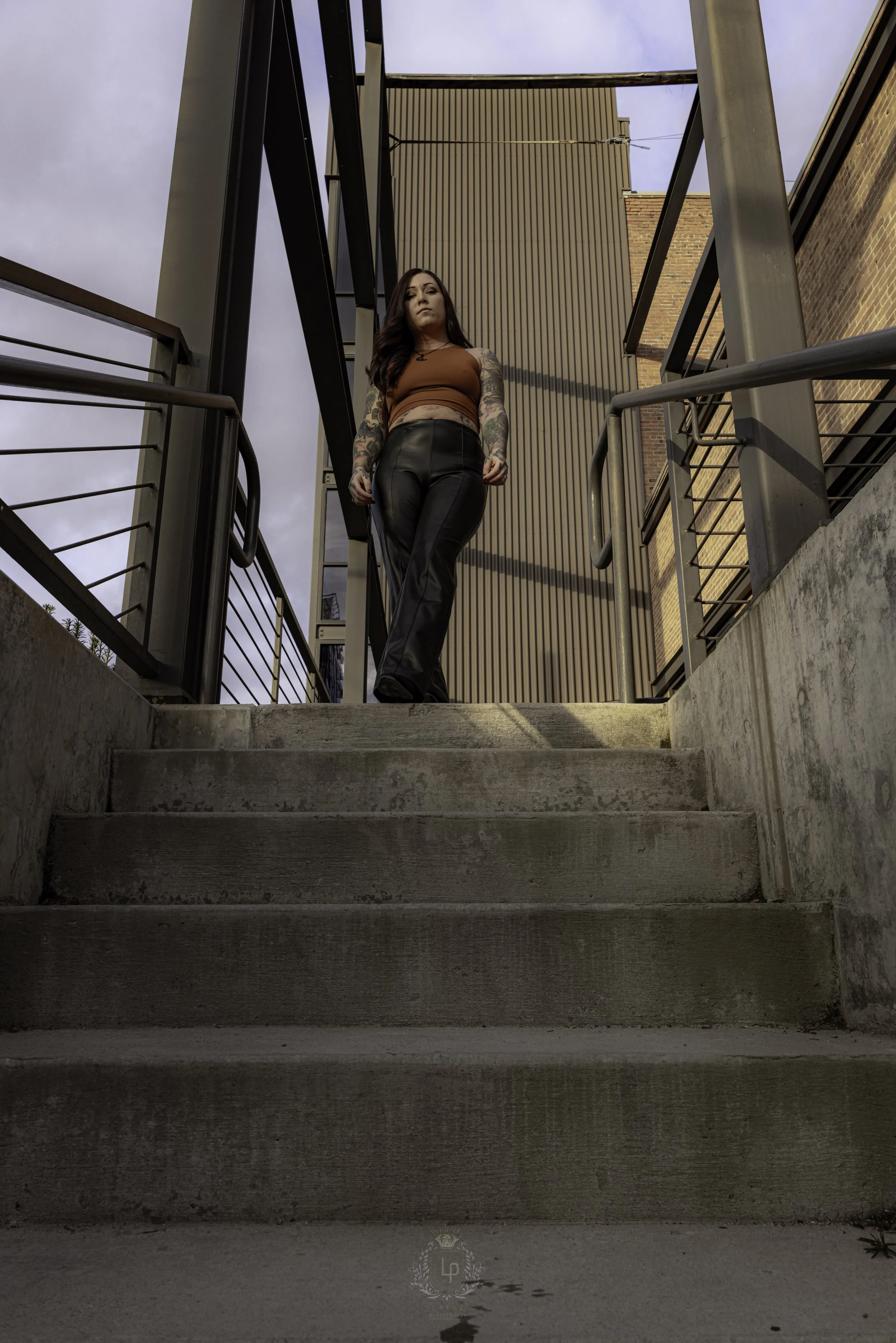 Woman with tattoos wearing a brown crop top and black leather pants walking down concrete stairs on an outdoor staircase, with modern metal railing and building structures in the background, under a cloudy sky.