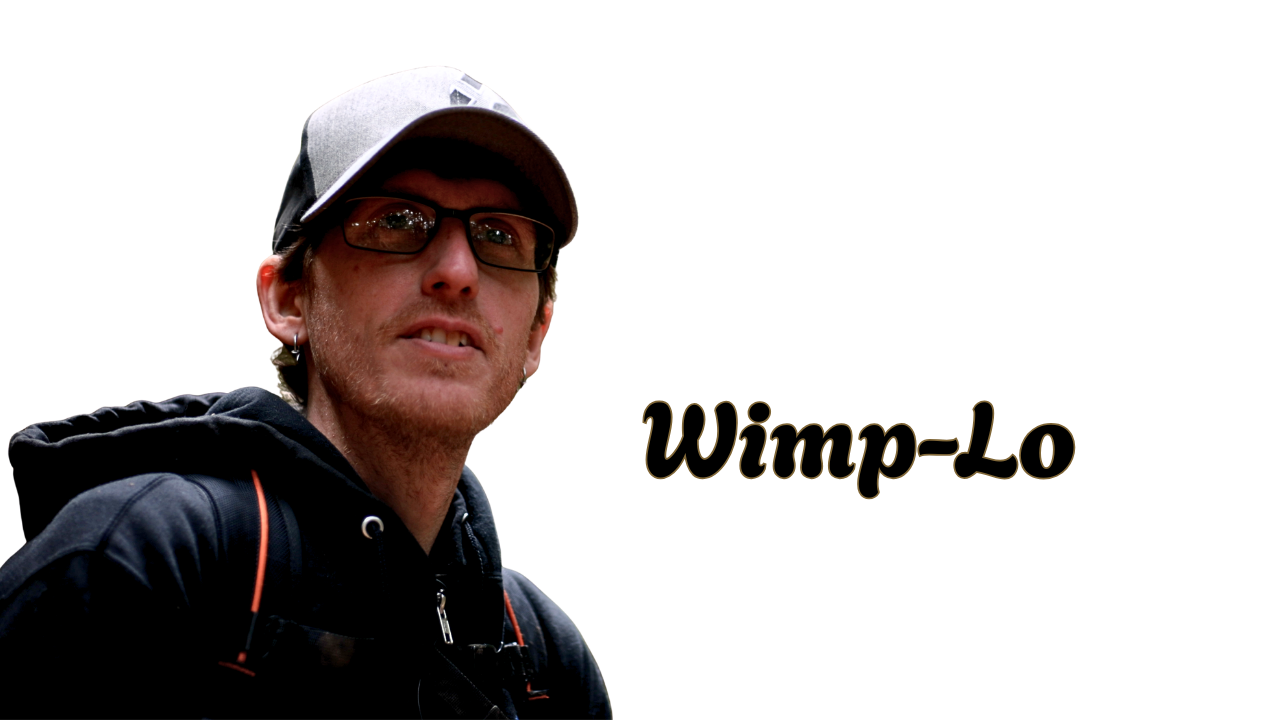 A man wearing glasses, a baseball cap, and a black hoodie, standing outdoors with a pixelated background. The text 'Wimp-Lo' is displayed next to him.