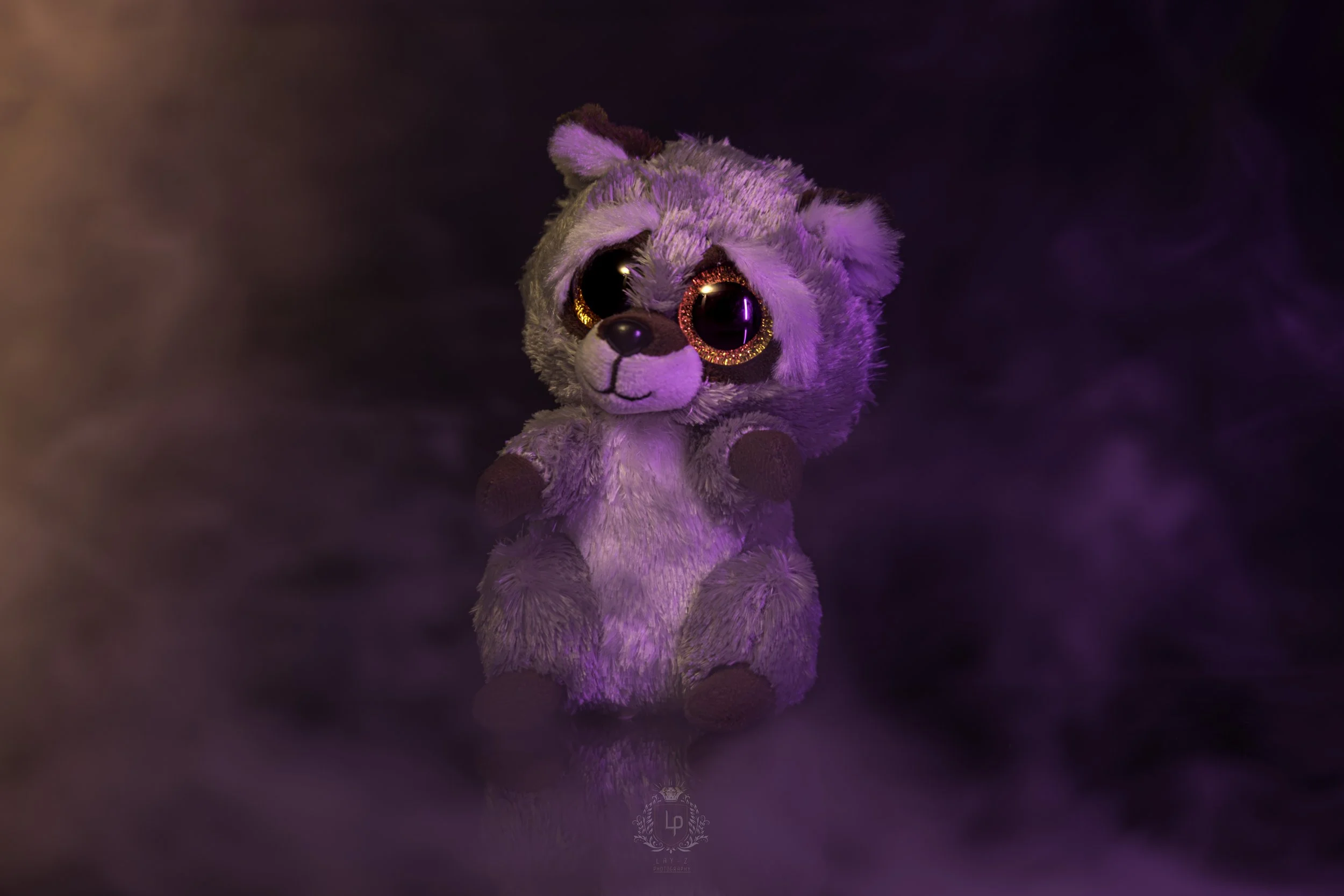 A plush teddy bear with large, shiny, black eyes accented with glittery pink rings, sitting against a purple misty background.