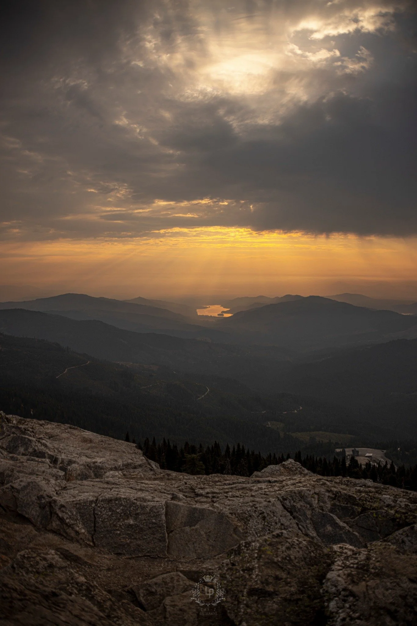 A scenic view of mountain ranges at sunset with clouds and rays of sunlight breaking through, overlooking a lake in the distance.