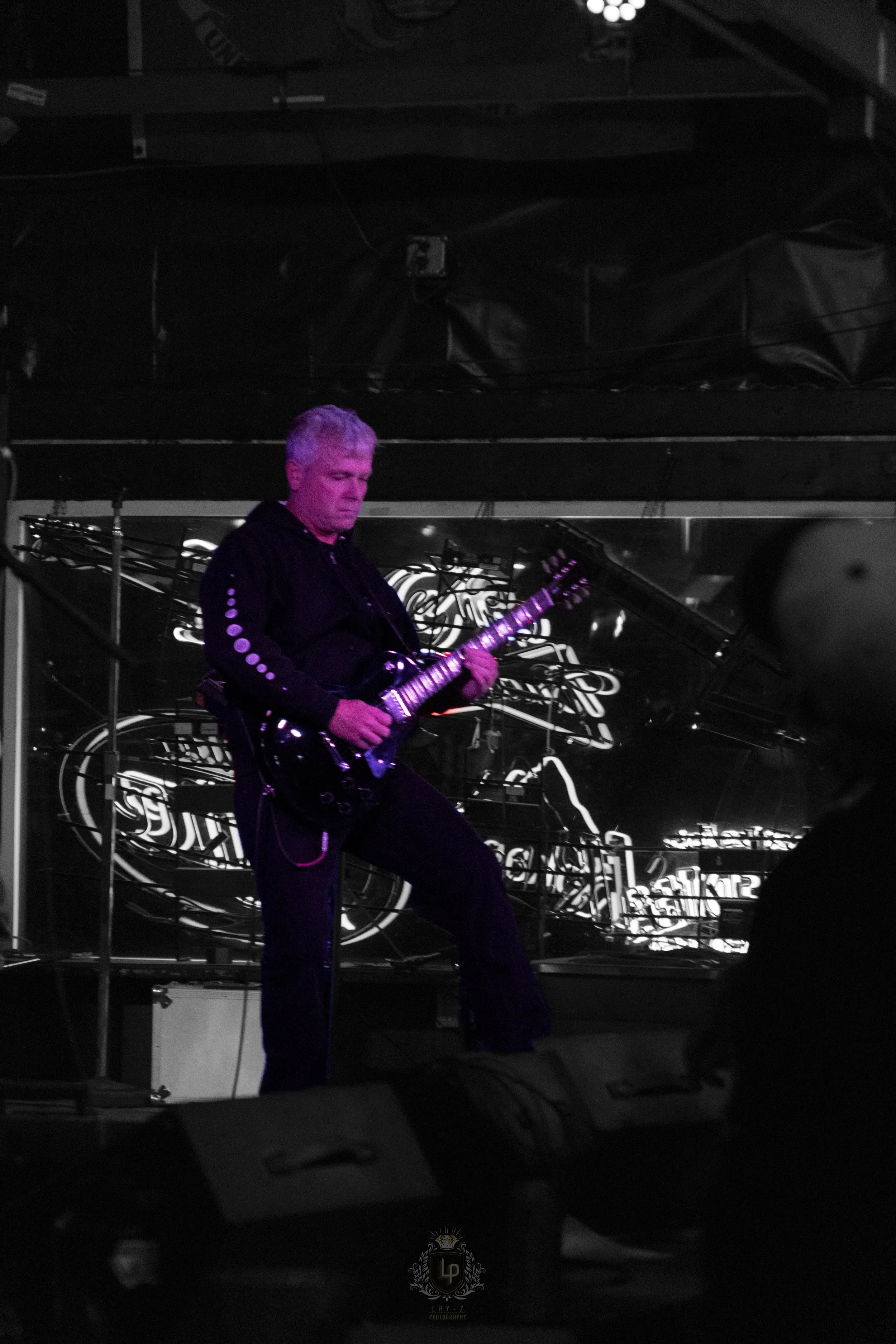 A man with gray hair is playing an electric guitar on stage with a dark background and bright stage lights.
