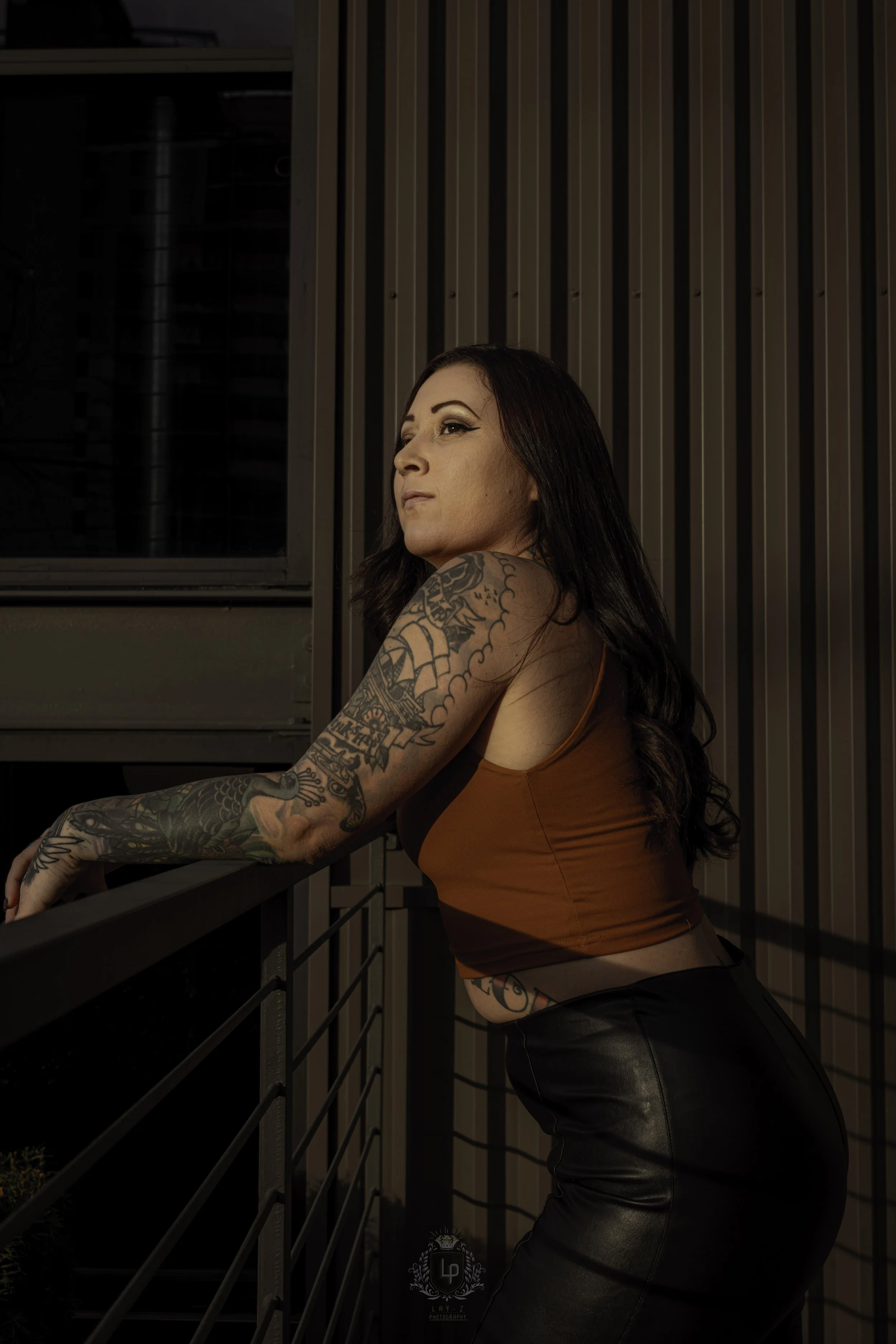 A woman with long dark hair and tattoos on her arm and side, wearing a brown sleeveless top and black leather pants, leaning on a balcony railing at night, looking to the side.