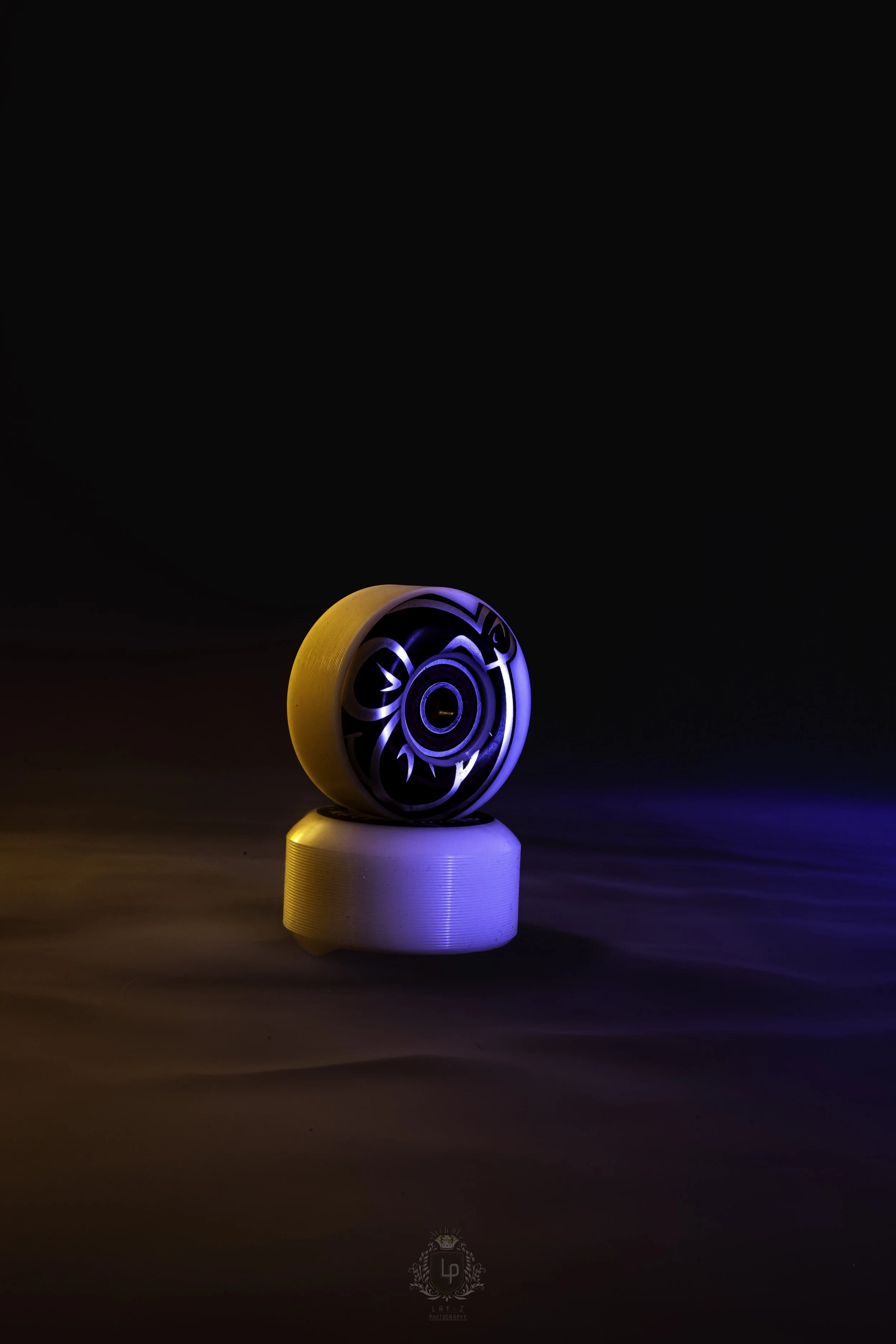 A black circular object with a stylized floral design on its surface, illuminated with blue and white lights, placed on a white cylindrical pedestal, against a dark background with multicolored light reflections.