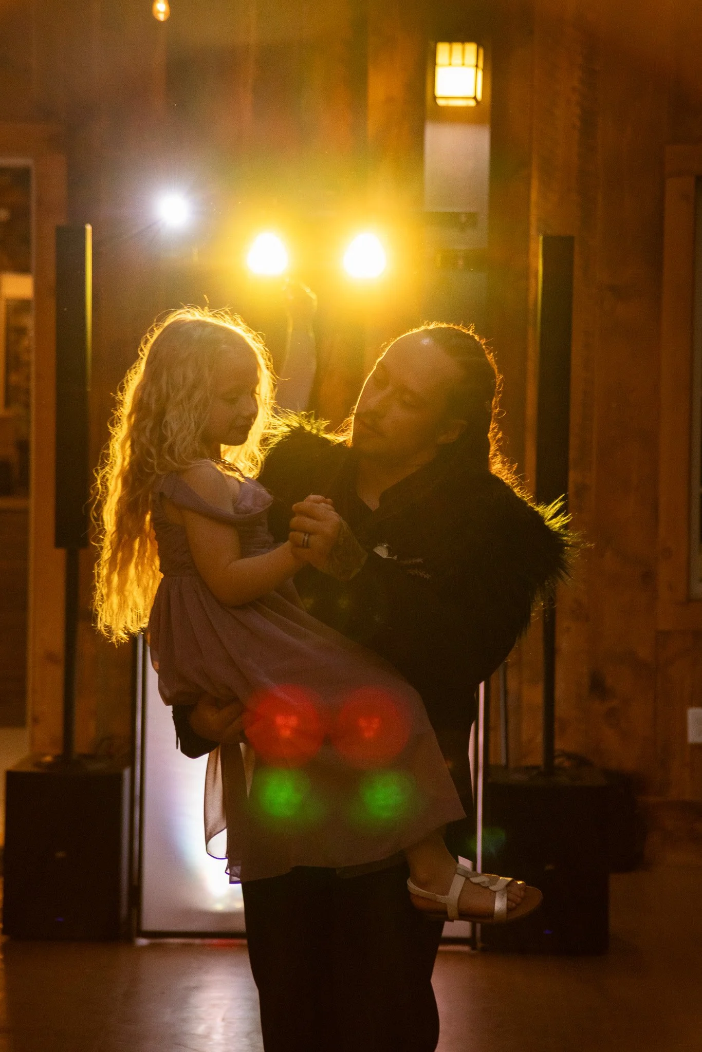 A man dancing with a young girl in a dimly lit room with warm yellow lighting and wooden walls.
