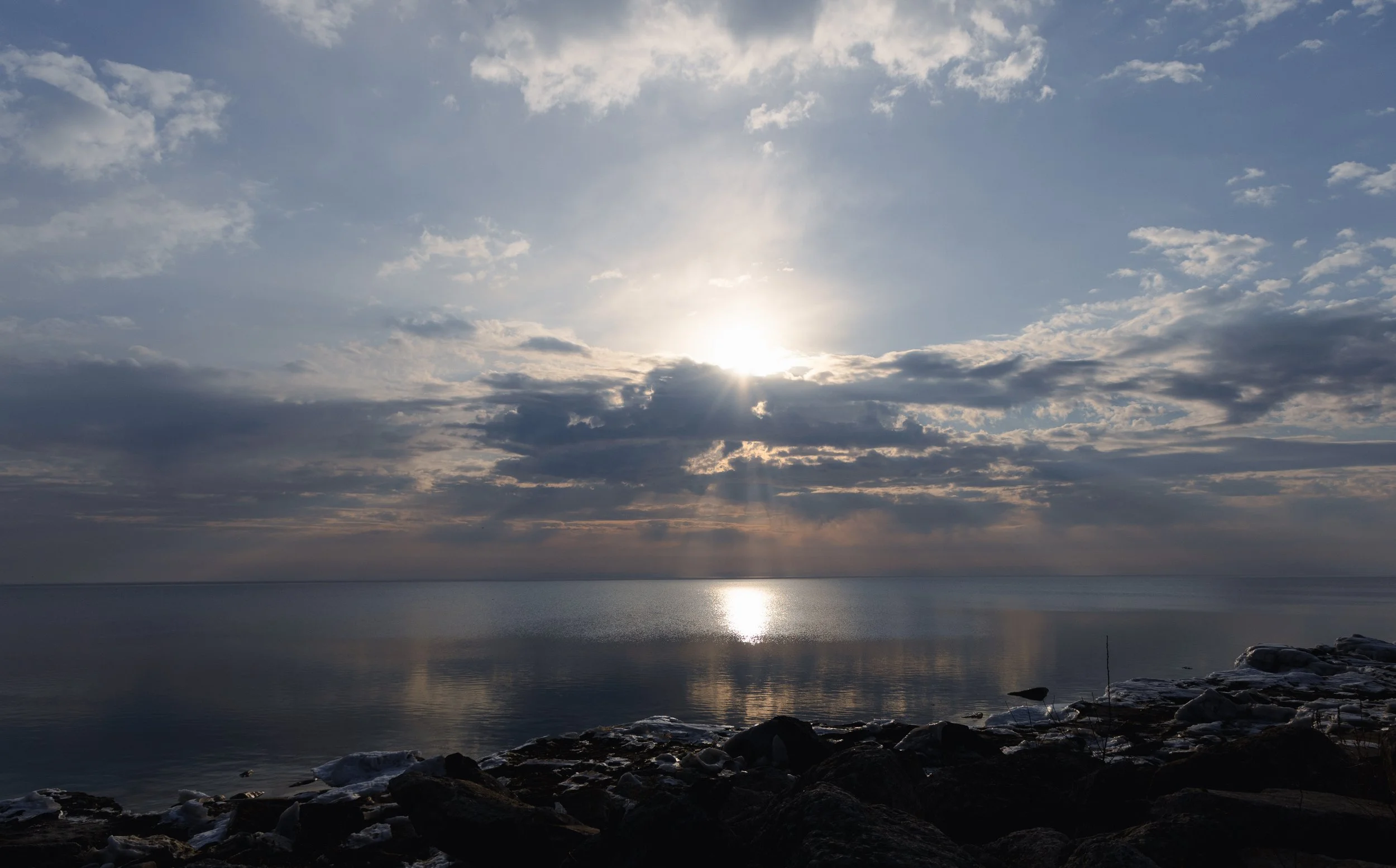Sun shining through clouds over a calm body of water with rocks along the shoreline.