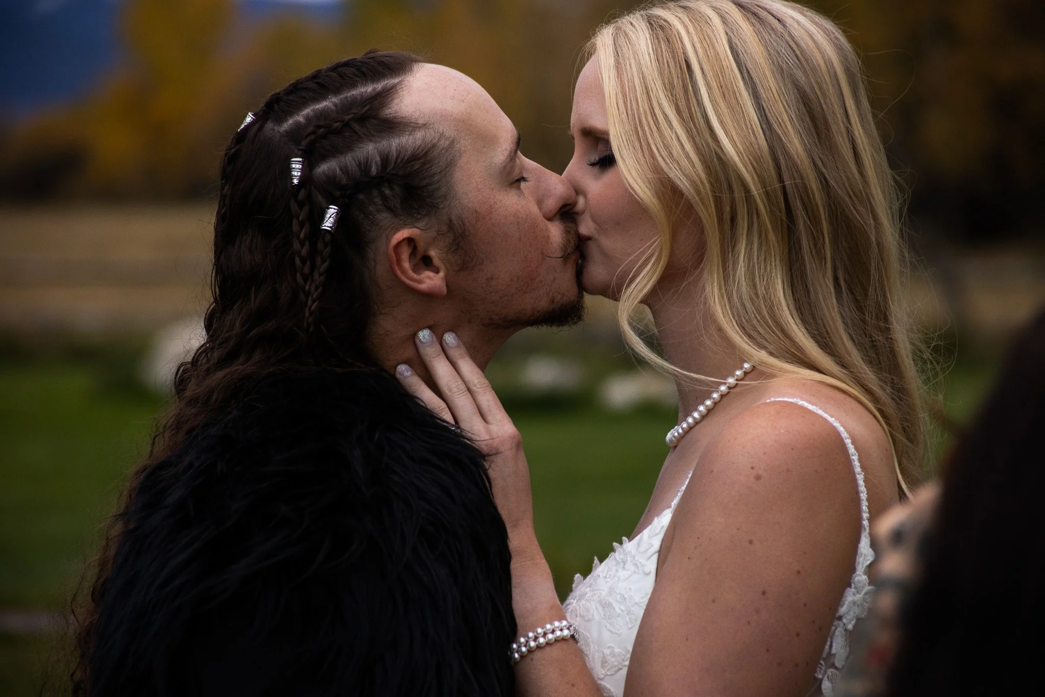 A couple sharing a kiss outdoors, the woman wearing a white lace dress, pearl jewelry, and the man with braided hair and a black fur coat, with trees and grass in the background.