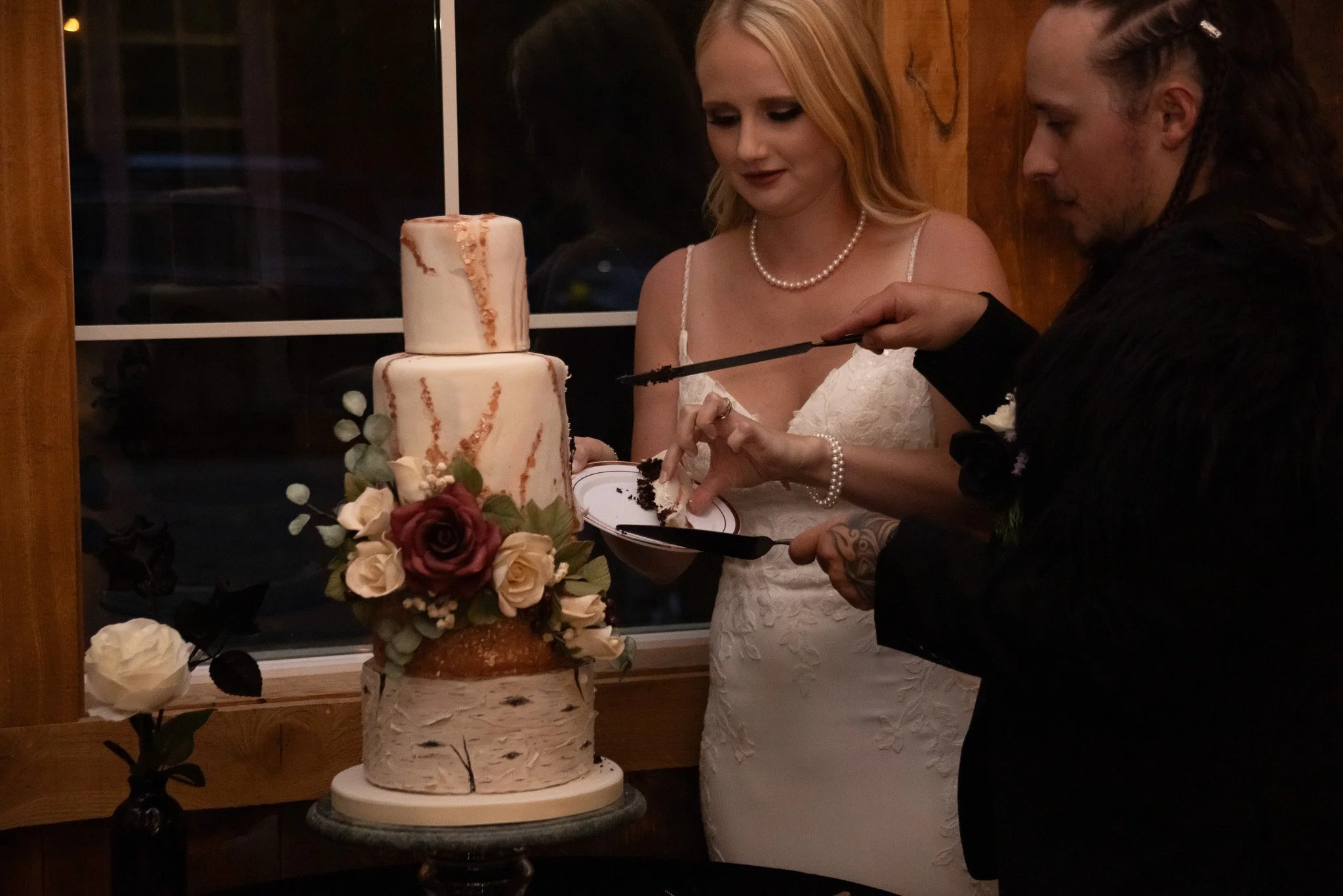 A bride and groom cut their wedding cake together. The cake has two tiers with floral decorations and is placed on a stand. The bride is wearing a white lace dress and pearl jewelry, while the groom is dressed in black. They are indoors near a window