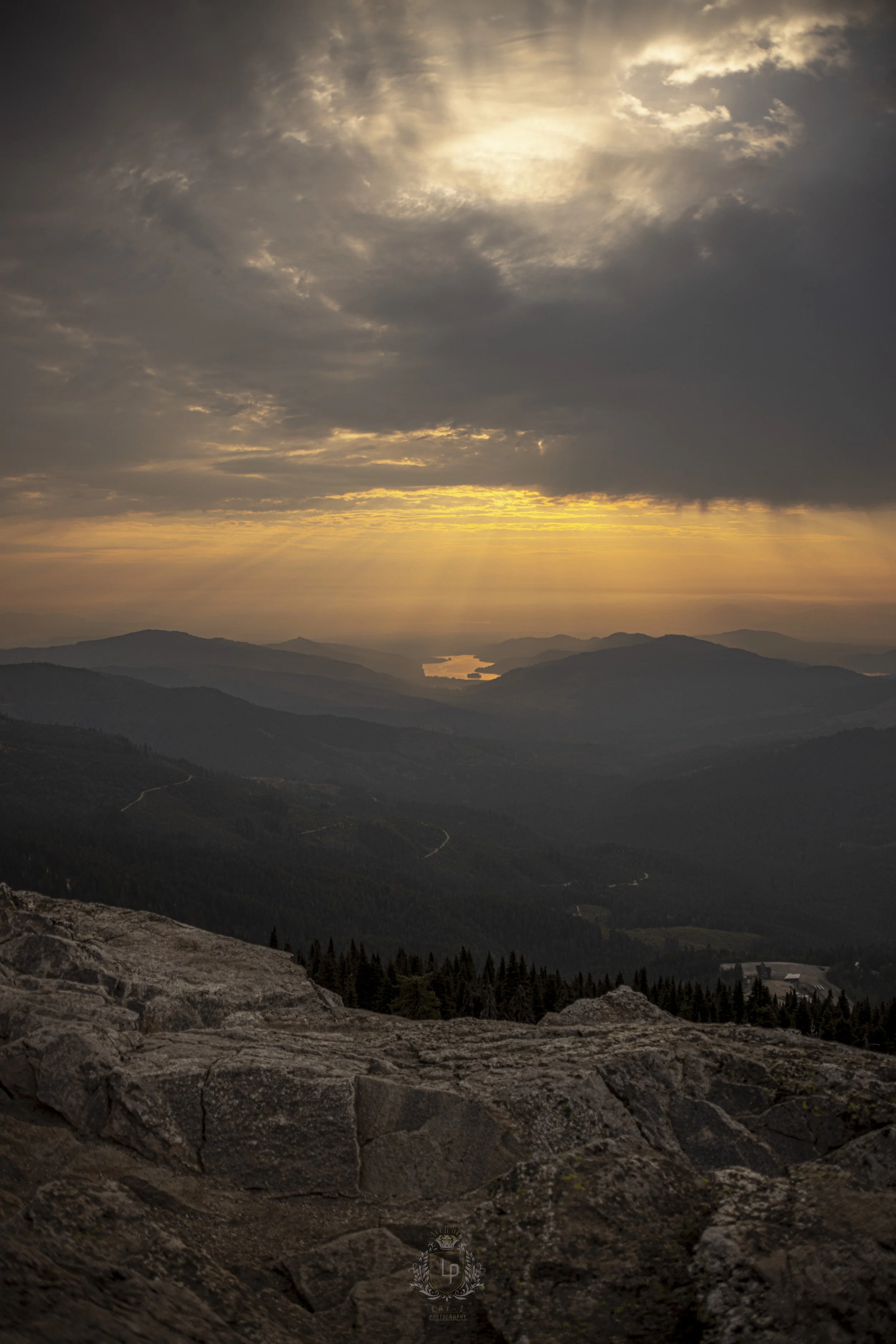 A scenic mountain landscape during sunset with layered mountain ridges, a winding road, a lake, and a sky with clouds and sun rays.