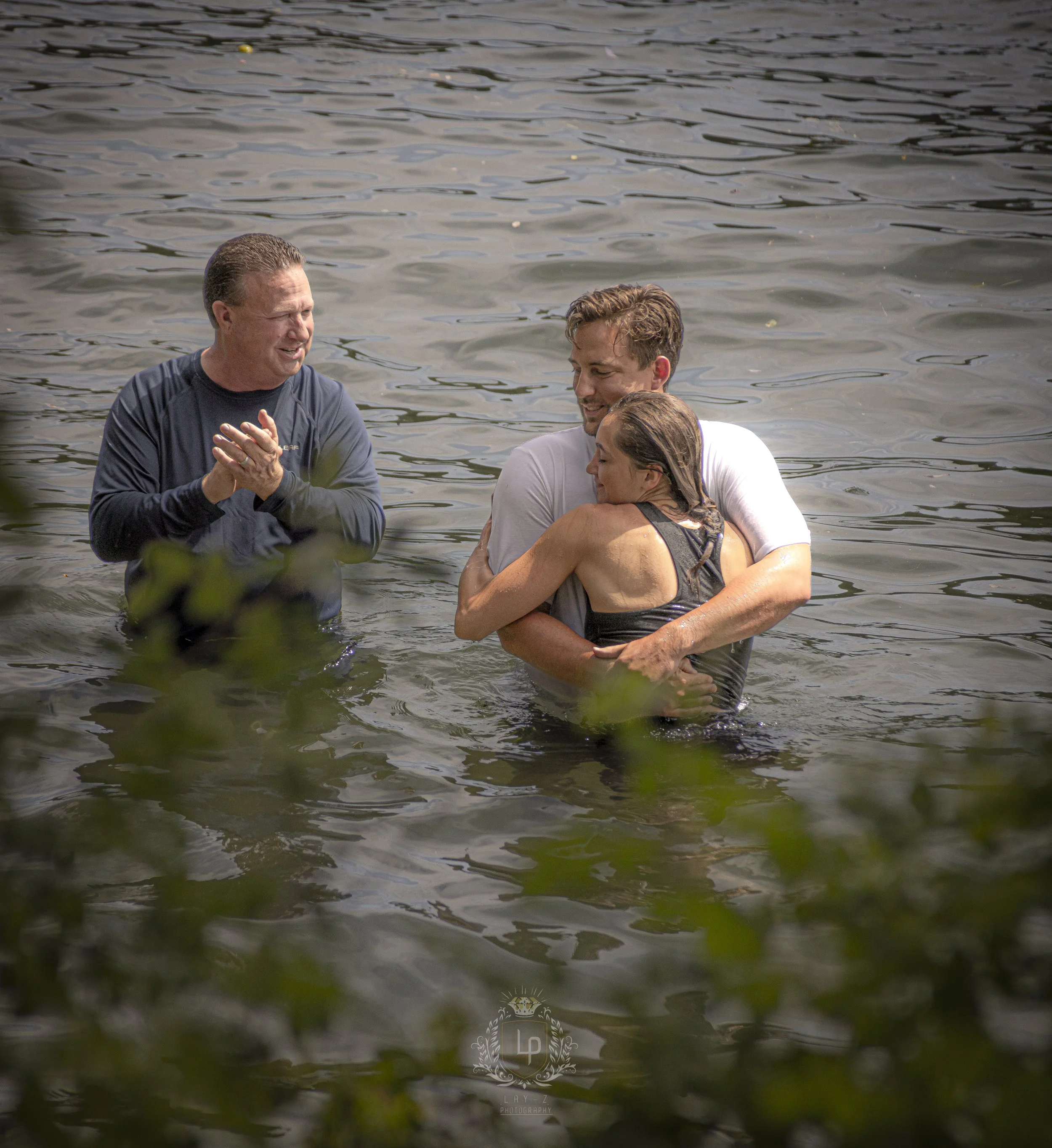 A couple is embracing in a body of water with a man standing nearby, clapping and smiling.