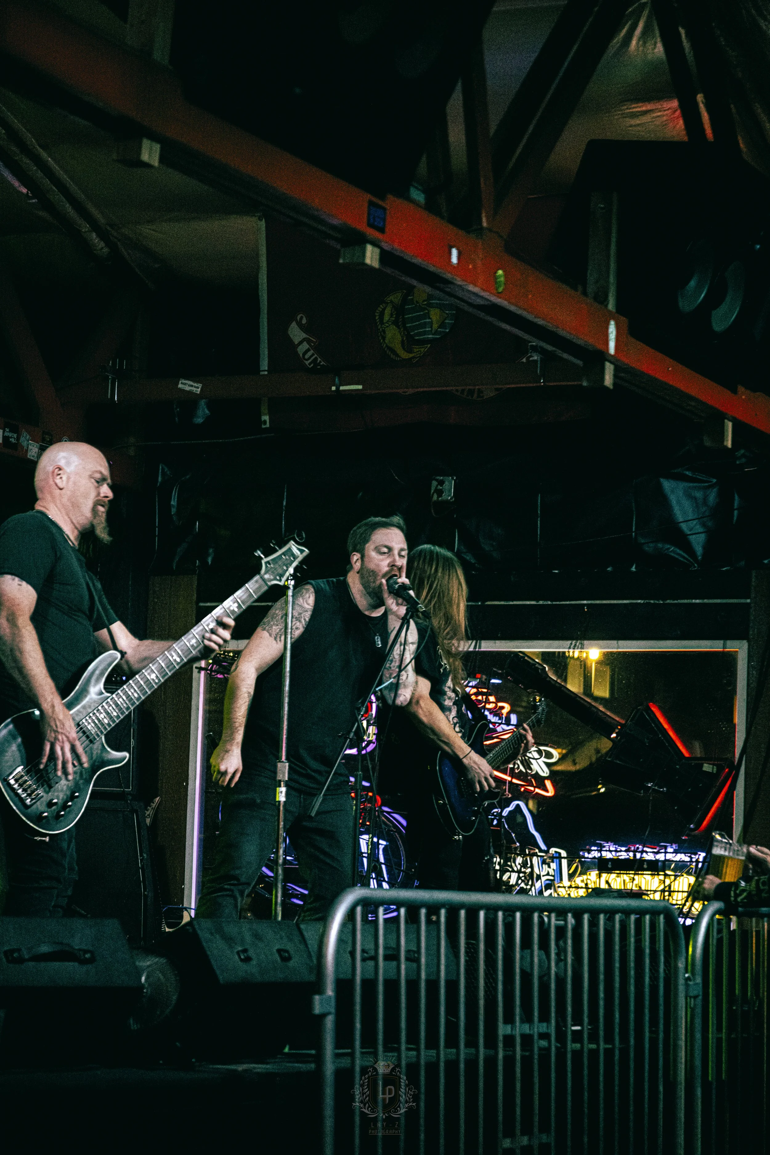 A live band performing on stage at a bar or music venue, with three musicians visible: a man playing bass guitar on the left, a vocalist with tattoos and long hair in the center singing into a microphone, and a woman playing electric guitar on the ri