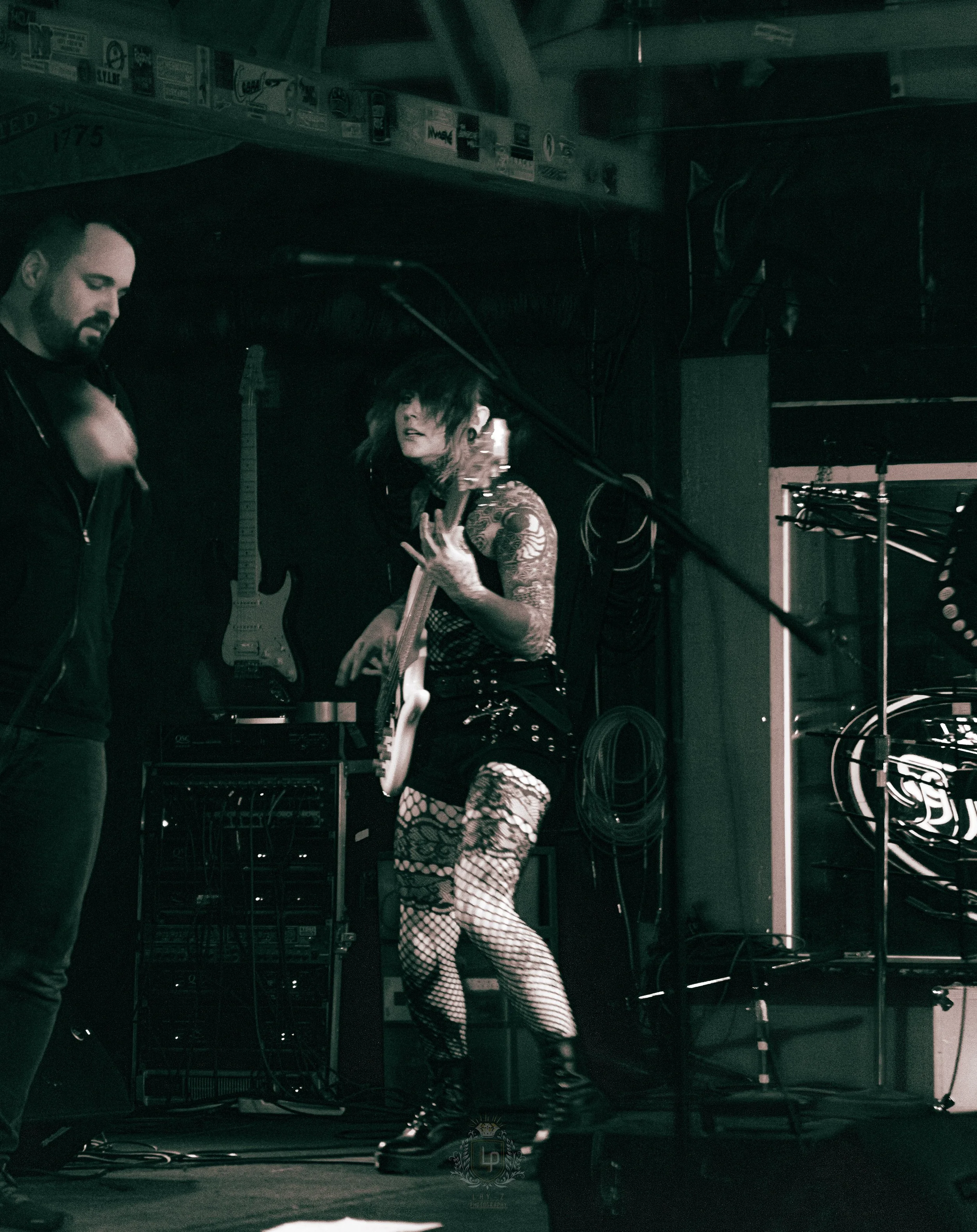 Female guitarist with tattoos, fishnet stockings, and combat boots performing on stage with a male bandmate at a music venue.