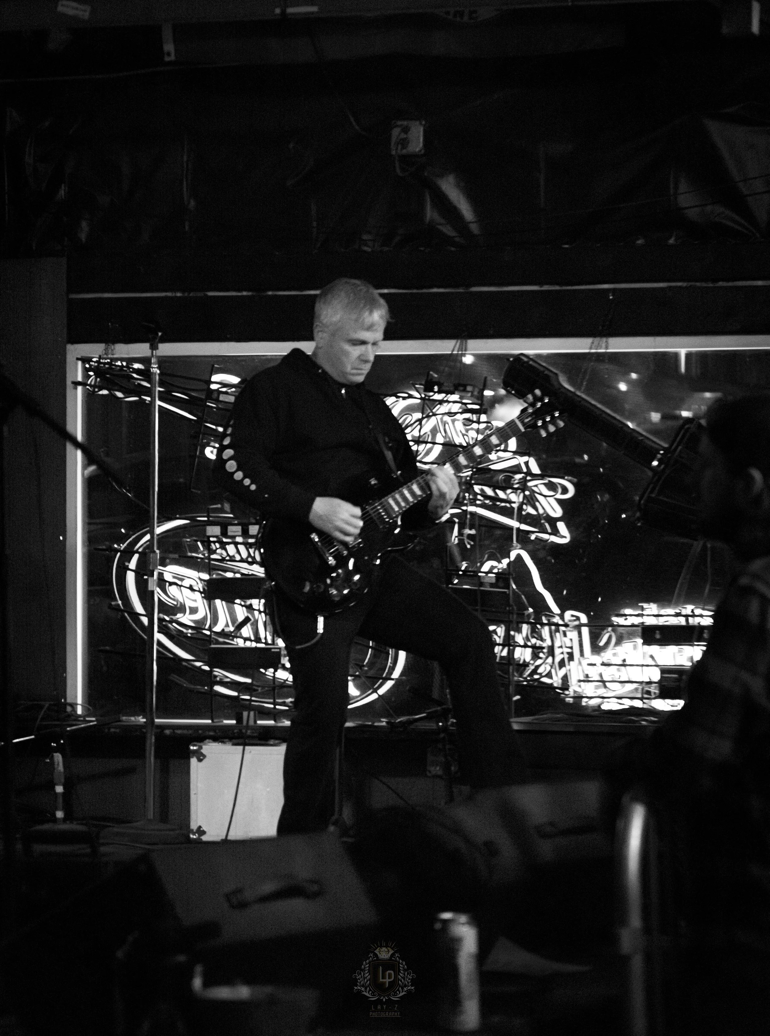A black and white photo of a man playing an electric guitar on stage, with neon lights in the background.