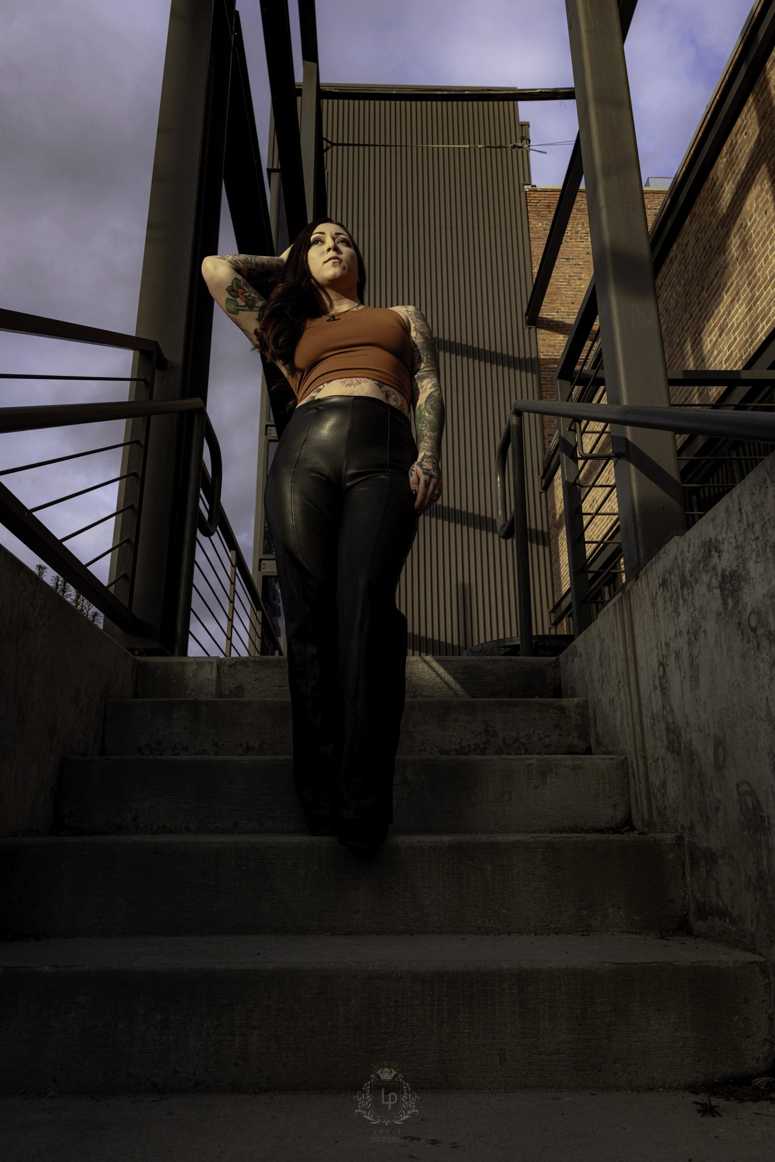 A woman with long dark hair and tattoos wearing a brown crop top and black leather pants standing on outdoor stairs with urban buildings and cloudy sky in the background.