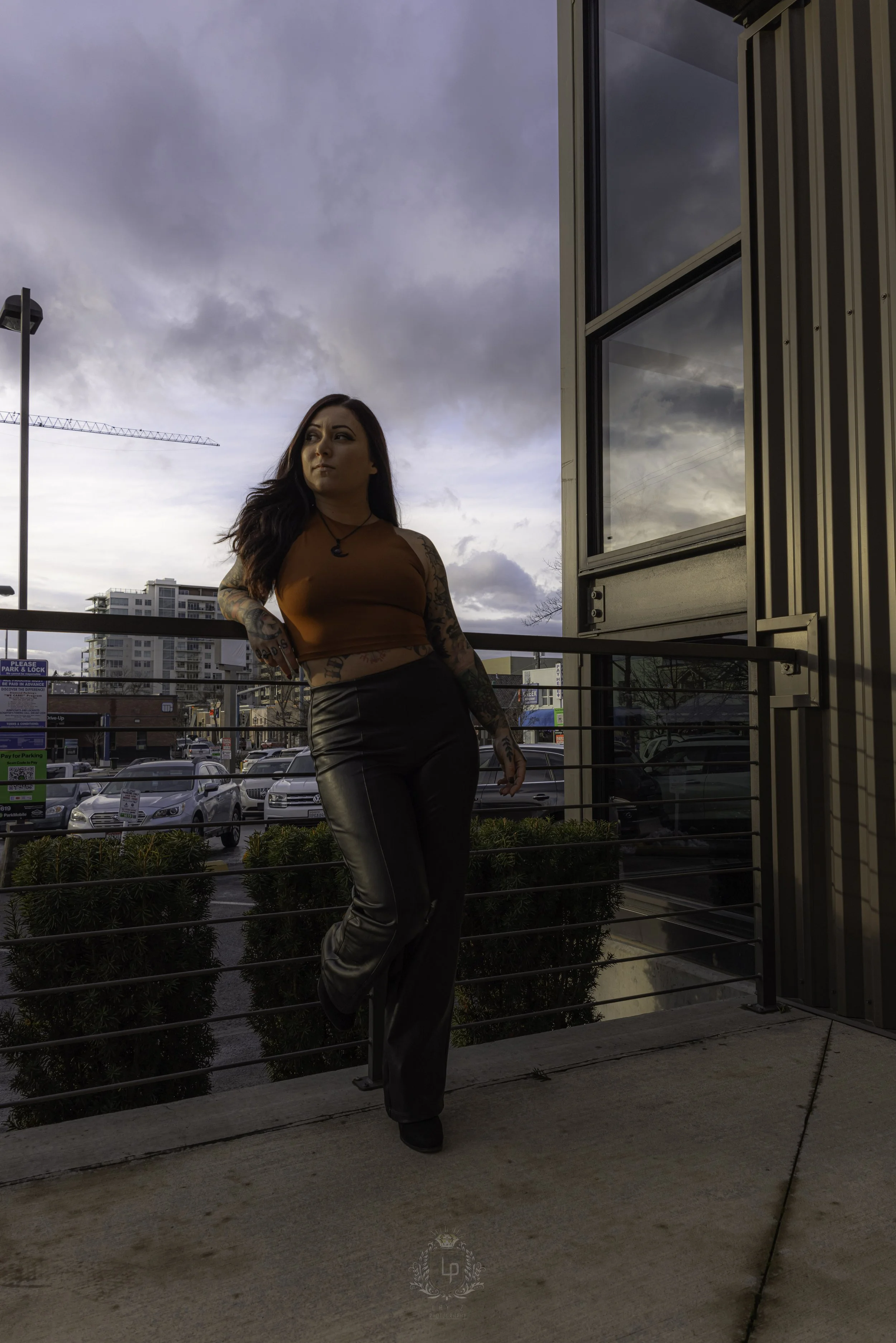 A woman with tattoos standing on a balcony, looking to the side, with a cloudy sky and city buildings in the background.