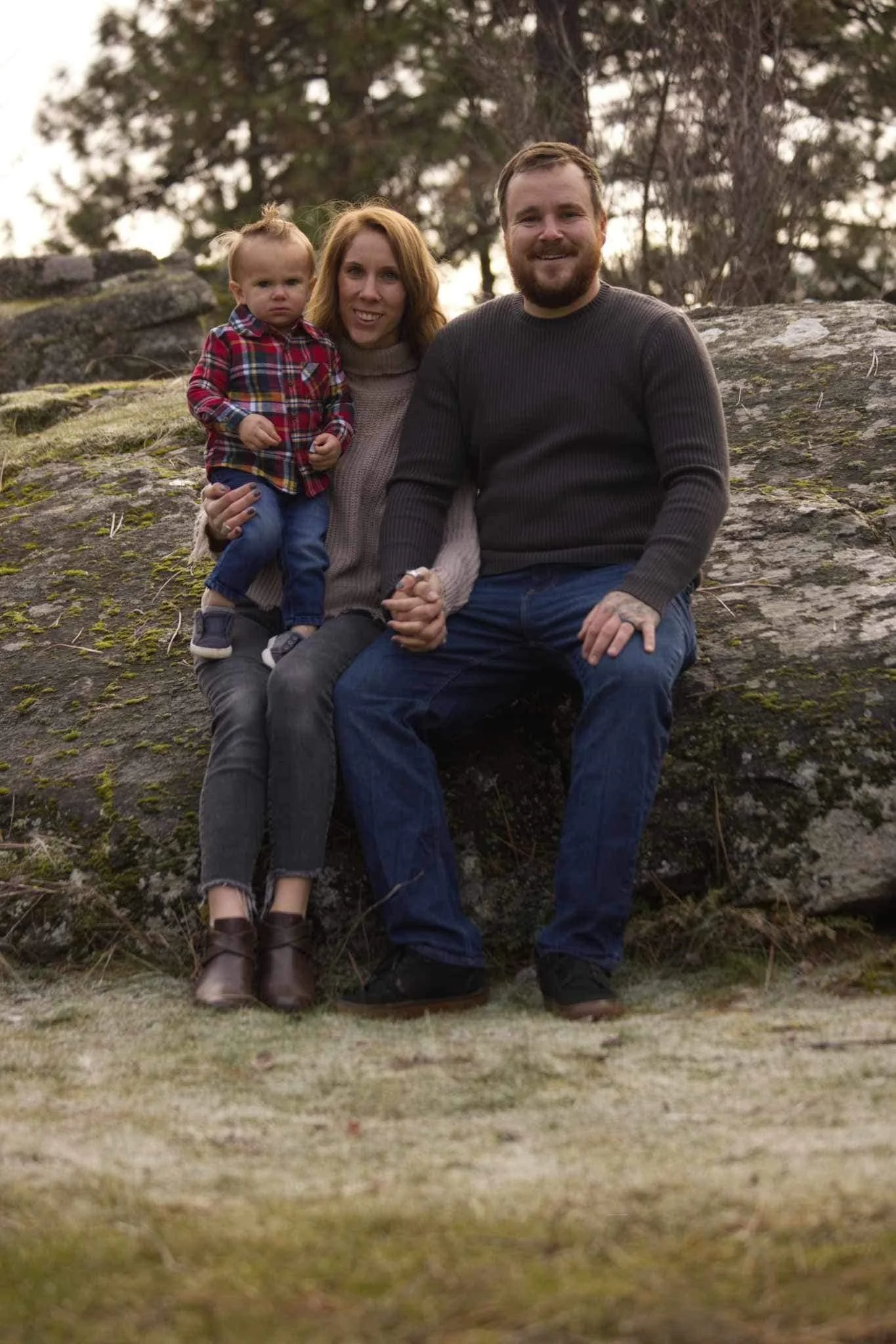 A family of three sitting on a large rock outdoors. The woman is holding a small child, and the man is sitting next to them, holding the woman's hand. The background features trees and a cloudy sky.