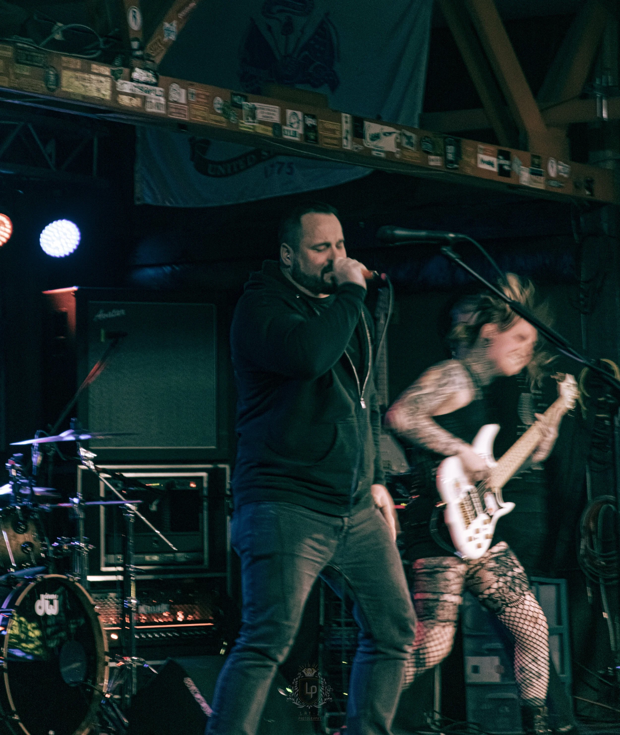 A band performs on stage, featuring a male vocalist with a microphone and a female guitarist with tattoos and fishnet stockings, in a dimly lit venue with music equipment and a flag in the background.