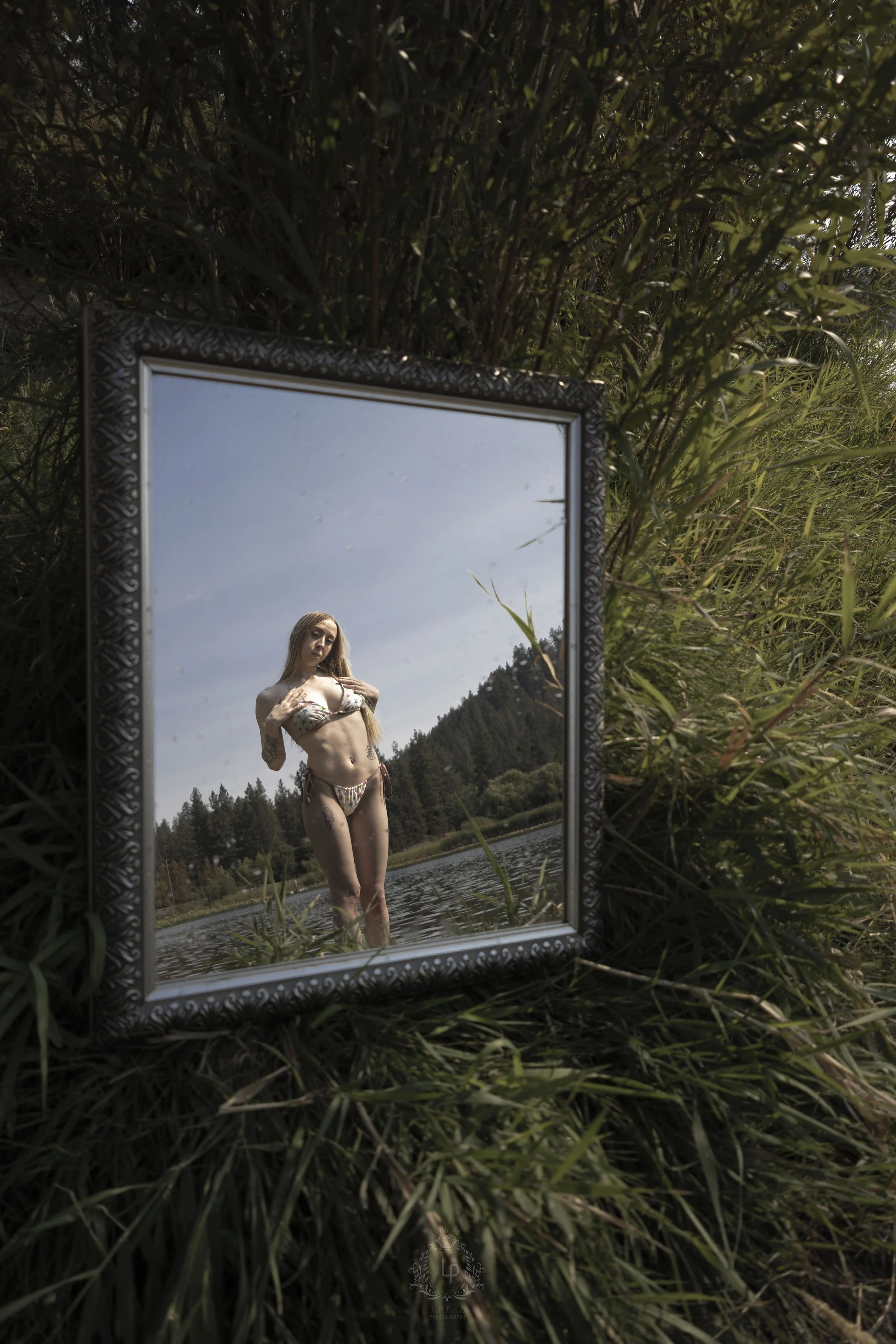 A mirror placed on the grass reflects a woman in a bikini standing in a lake with a forested hill in the background.