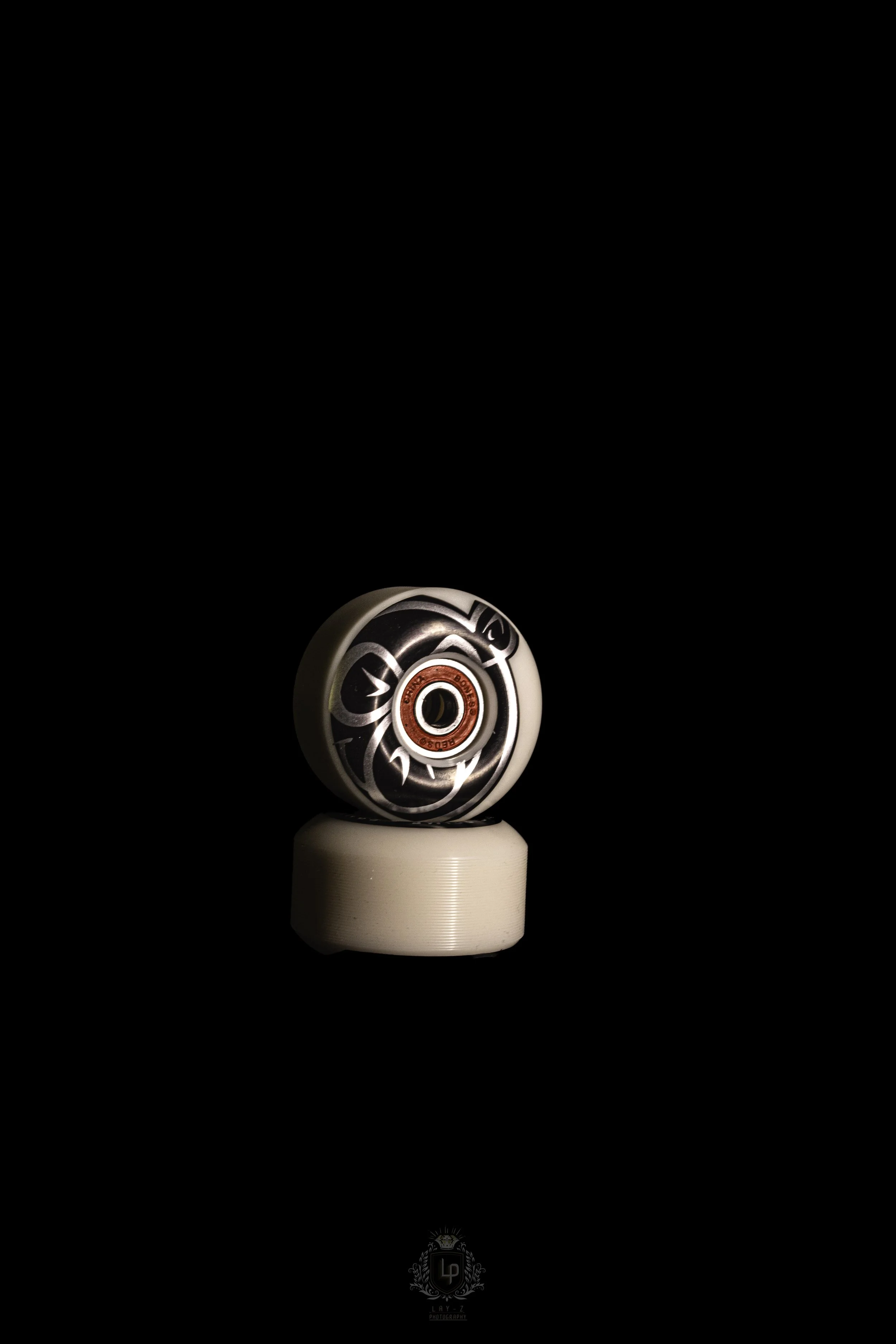 A skateboard wheel with black and white tribal design and red inner bearing sits on top of a plain white wheel against a black background.