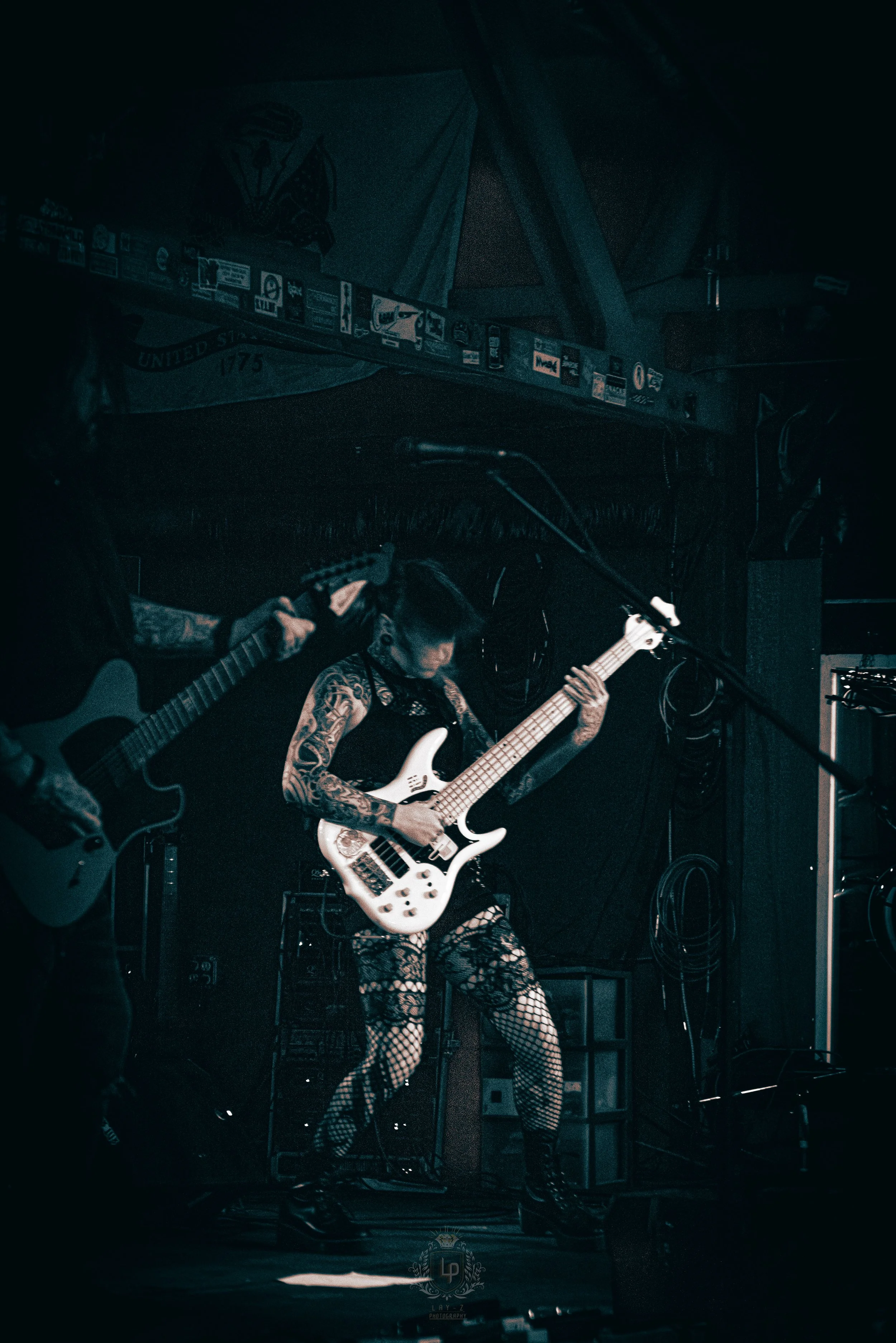 A female musician with tattoos wearing fishnet stockings, playing a white bass guitar on a dark stage, with a male guitarist on her left in a black shirt.