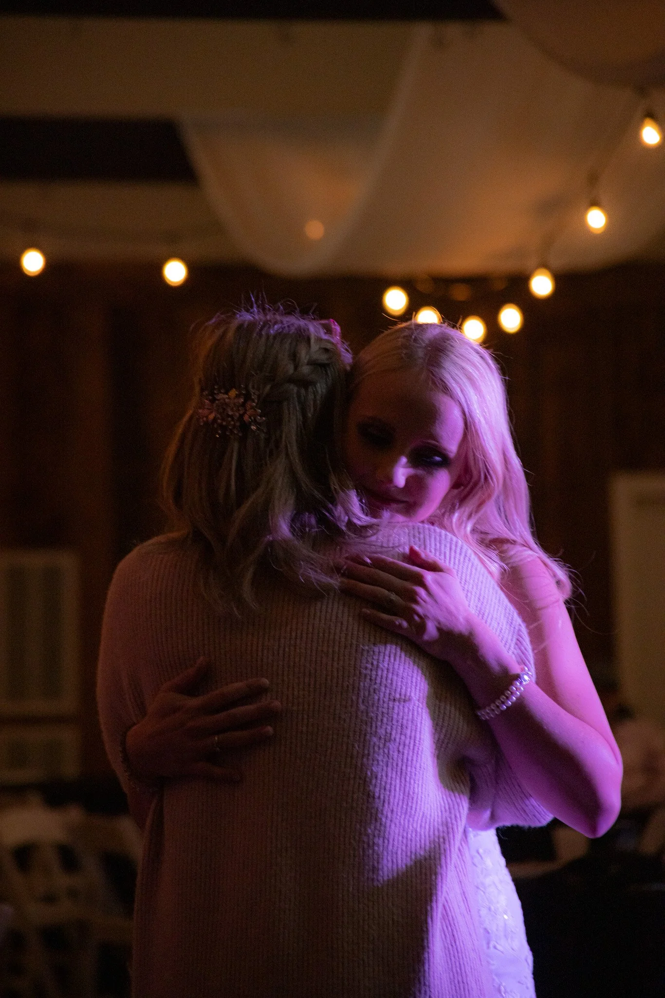 Two women hugging at a celebration or dance, with warm lighting and string lights overhead.