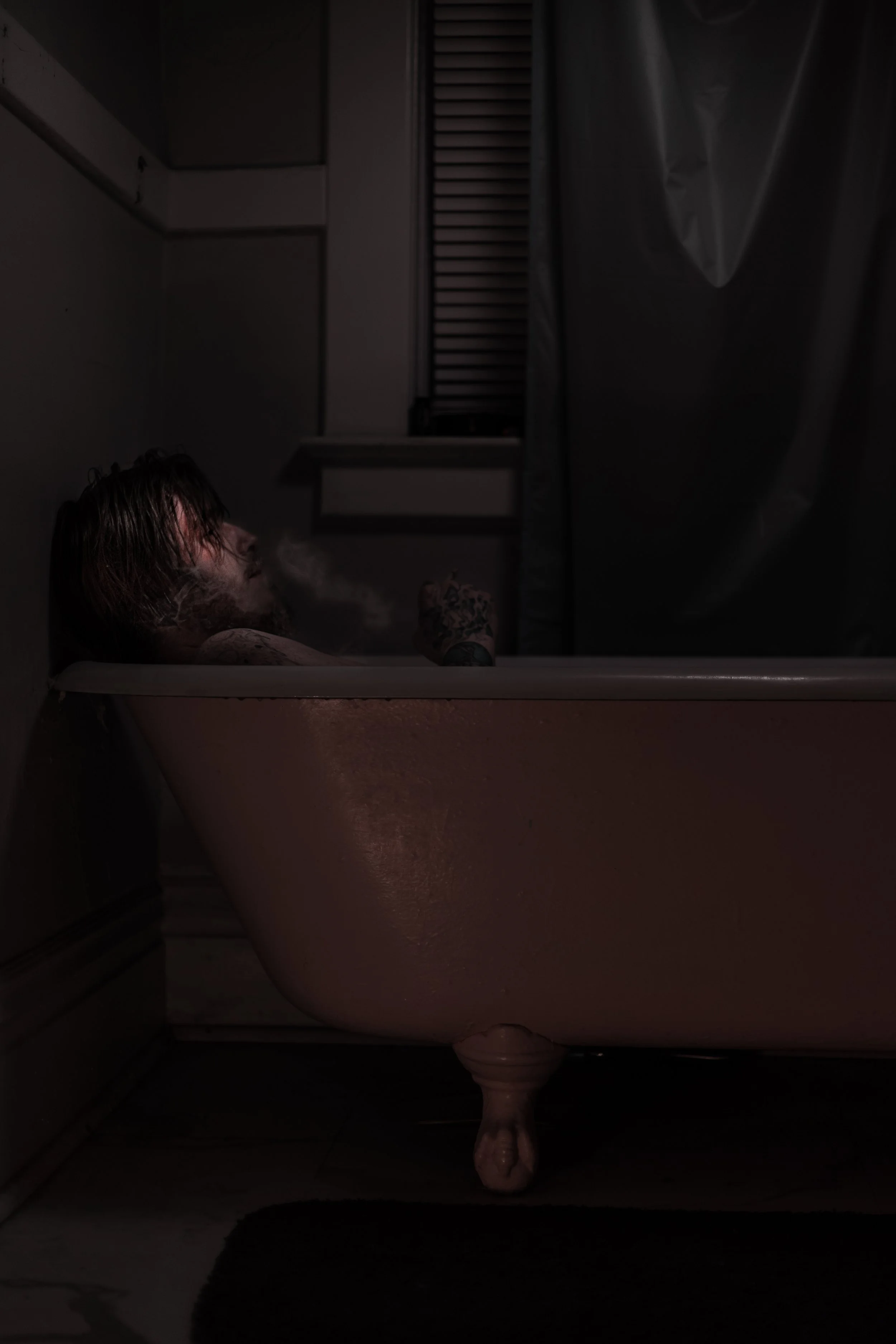 A man with long hair and tattoos is relaxing in a dimly lit vintage clawfoot bathtub, exhaling vapor in a dark bathroom.