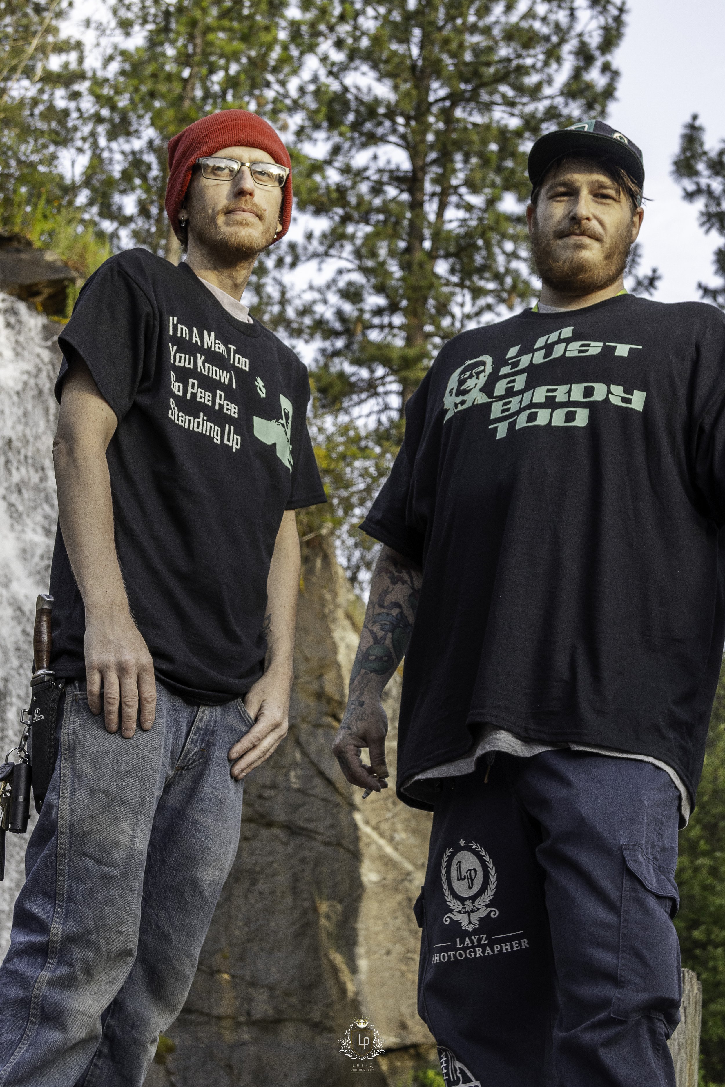 Two men standing outdoors near trees, wearing black T-shirts with printed text and graphics, one with a red beanie and glasses, the other with a black cap and tattoos on his arm.