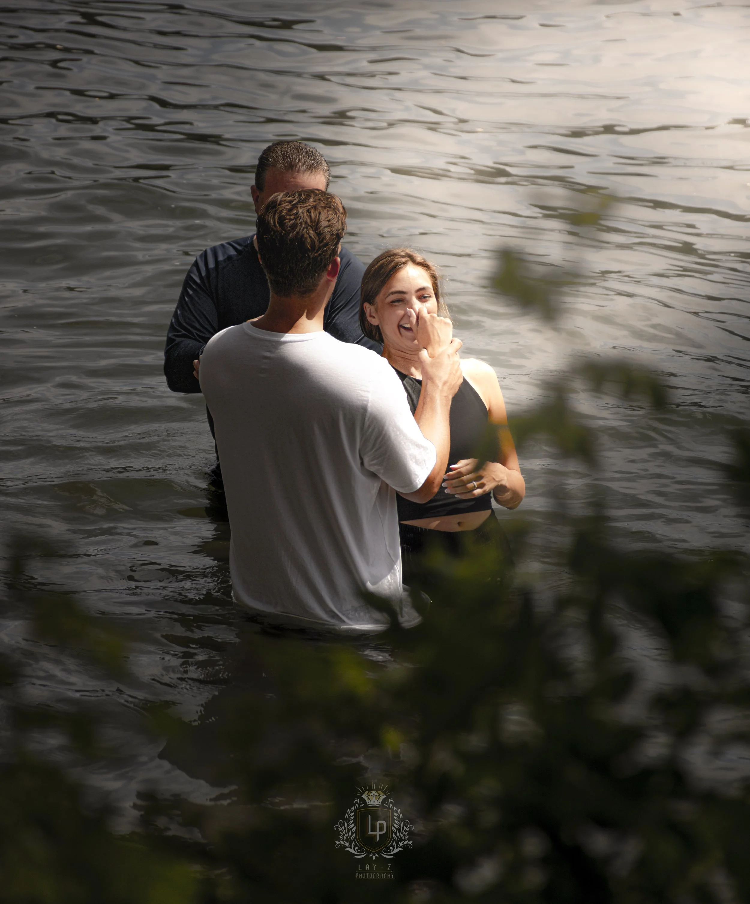 A woman is being baptized in a body of water while two men assist her, one holding her nose and the other supporting her back, during a sunny day.