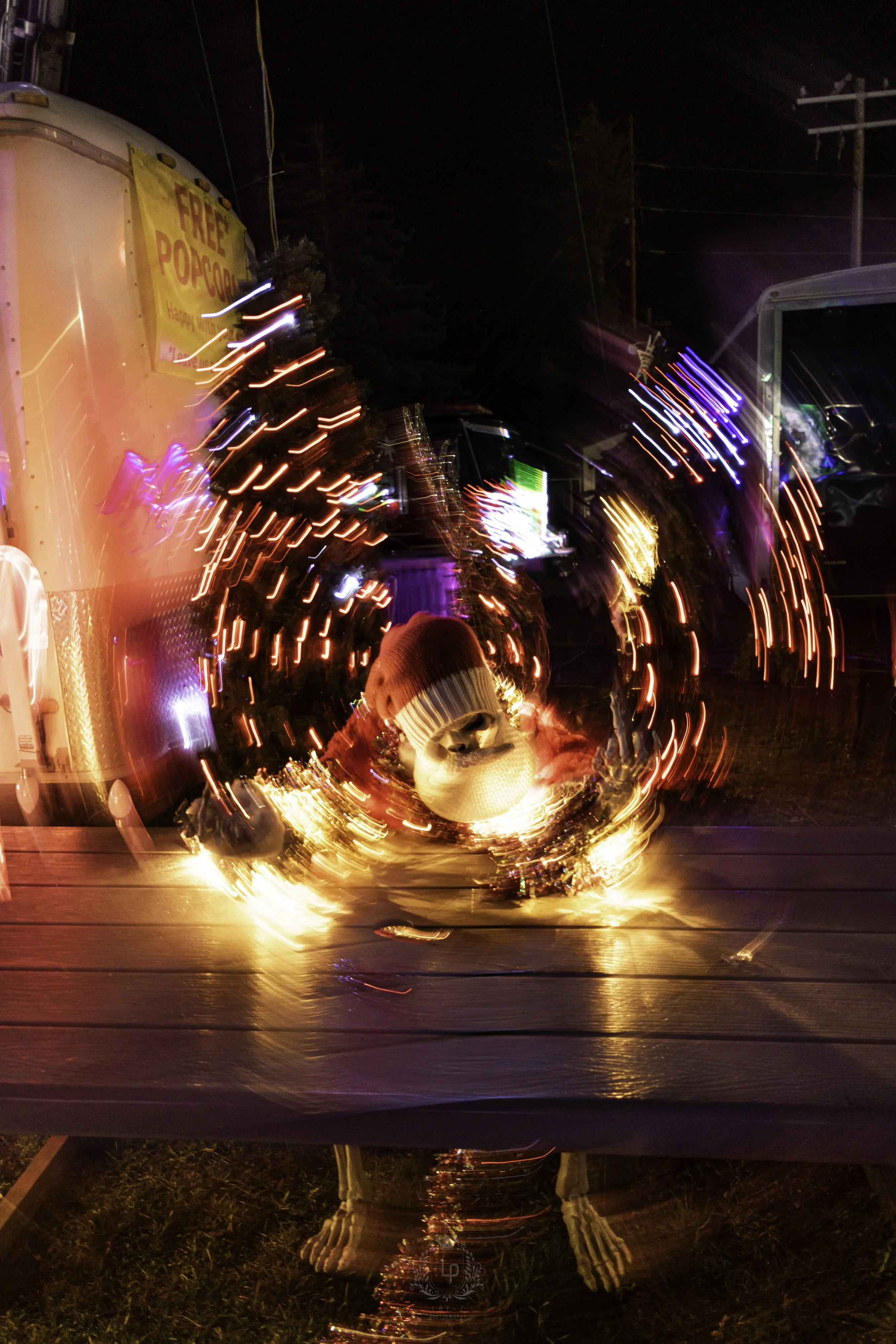 A skeleton wearing a red beanie and sunglasses, illuminated by colorful lights, is sitting outdoors at night at a table with a pumpkin and chewed flowers. In the background, there are food trucks and string lights.