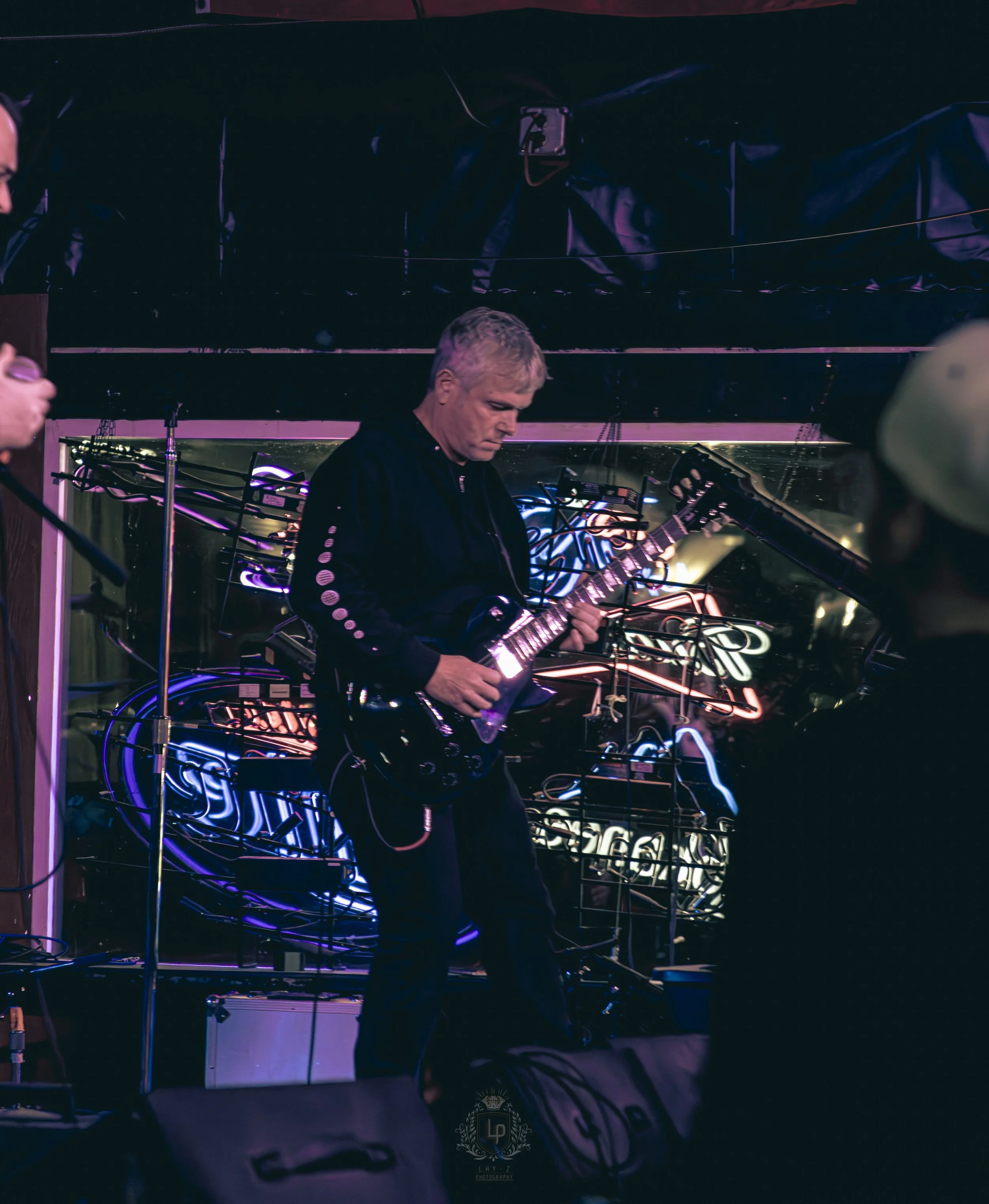 A man with blond hair playing an electric guitar on stage with colorful neon lights and photos in the background.