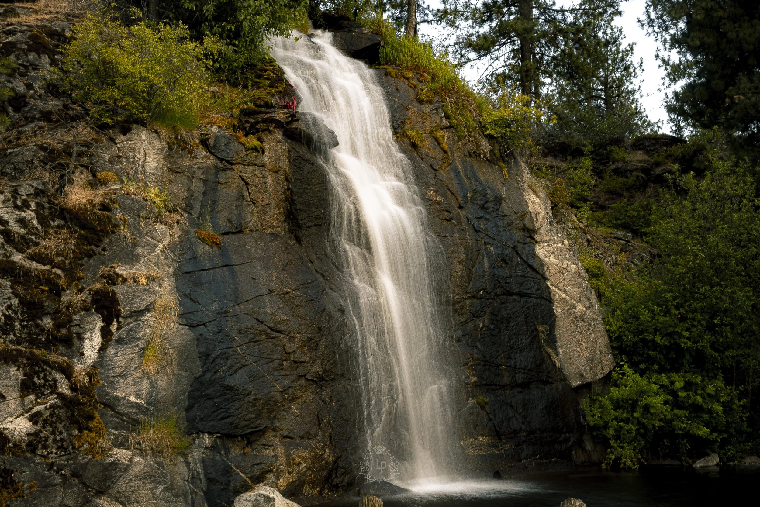 A scenic waterfall flowing down a rocky cliff surrounded by green trees and foliage.