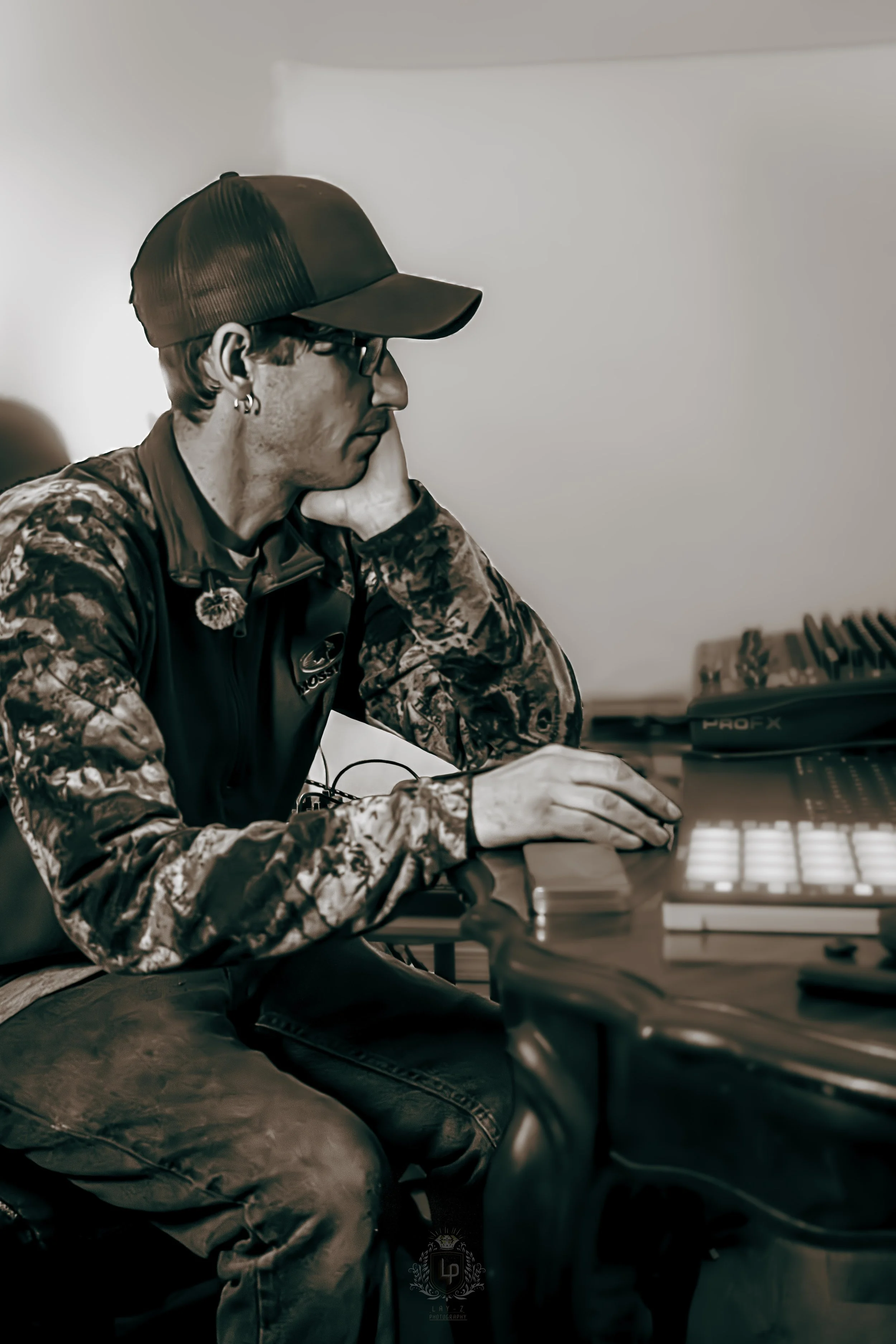 A young man wearing a camouflage jacket, black baseball cap, and glasses, sitting at a desk with his head resting on his hand, working on music production equipment.