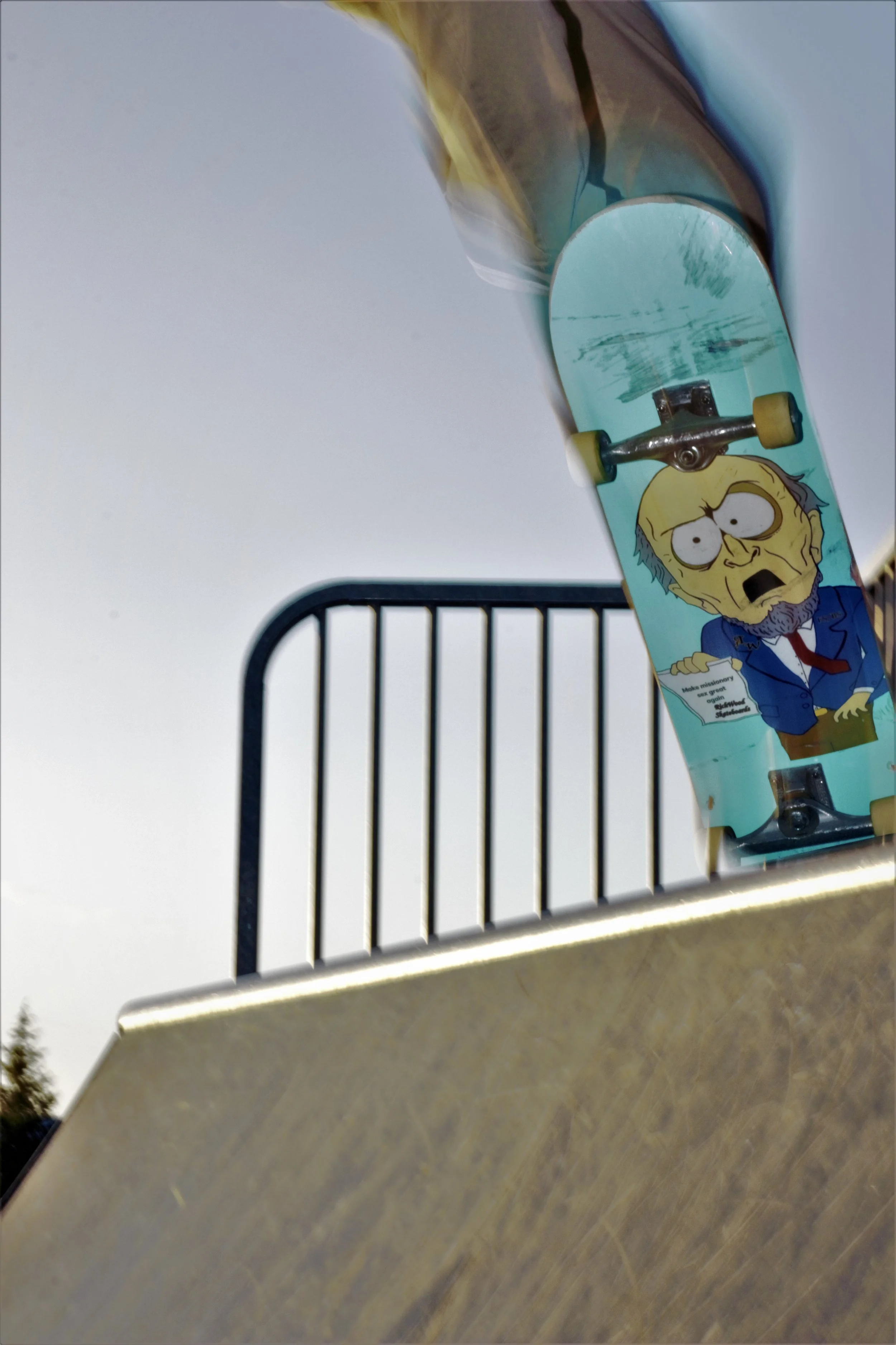 A skateboard with a cartoon character graphic leaning against a park structure. The cartoon character has a distressed expression, glasses, and is wearing a blue suit. The park structure has a metal railing and a metal surface.
