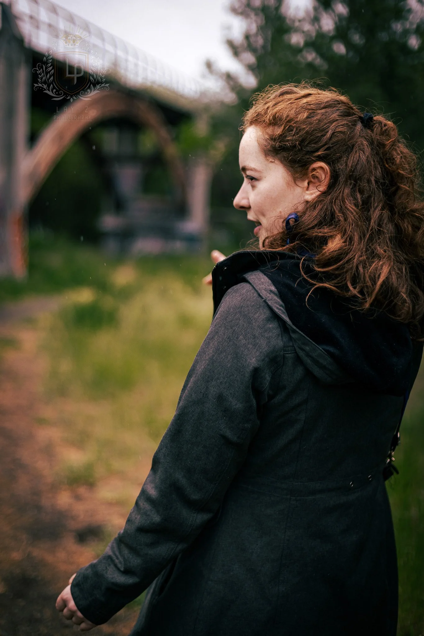 A woman with curly red hair wearing a dark jacket stands outdoors with greenery and a gazebo in the background.