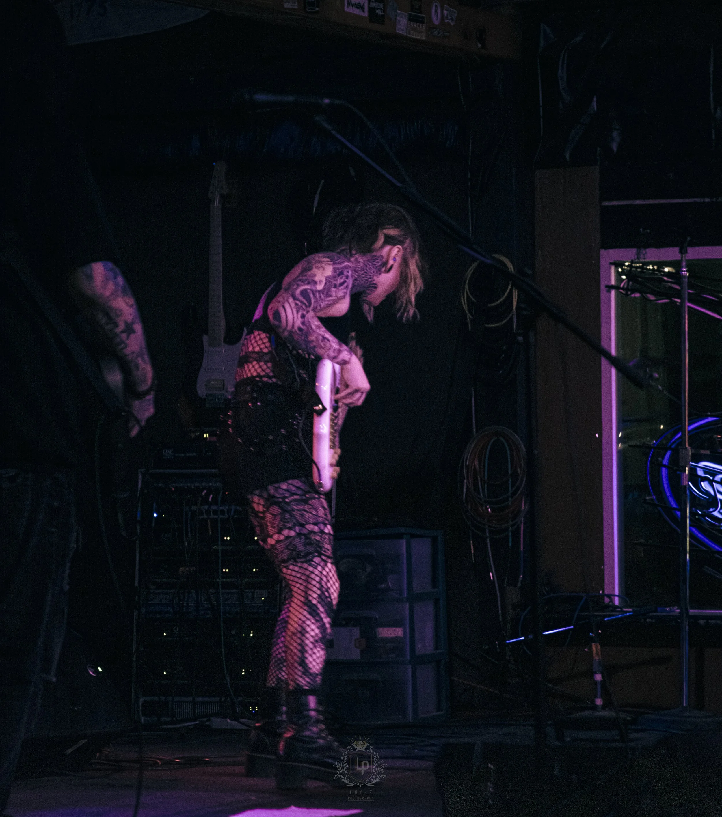 A person with tattoos and fishnet clothing on stage, holding an electric guitar, in a dark venue with purple and pink lighting.