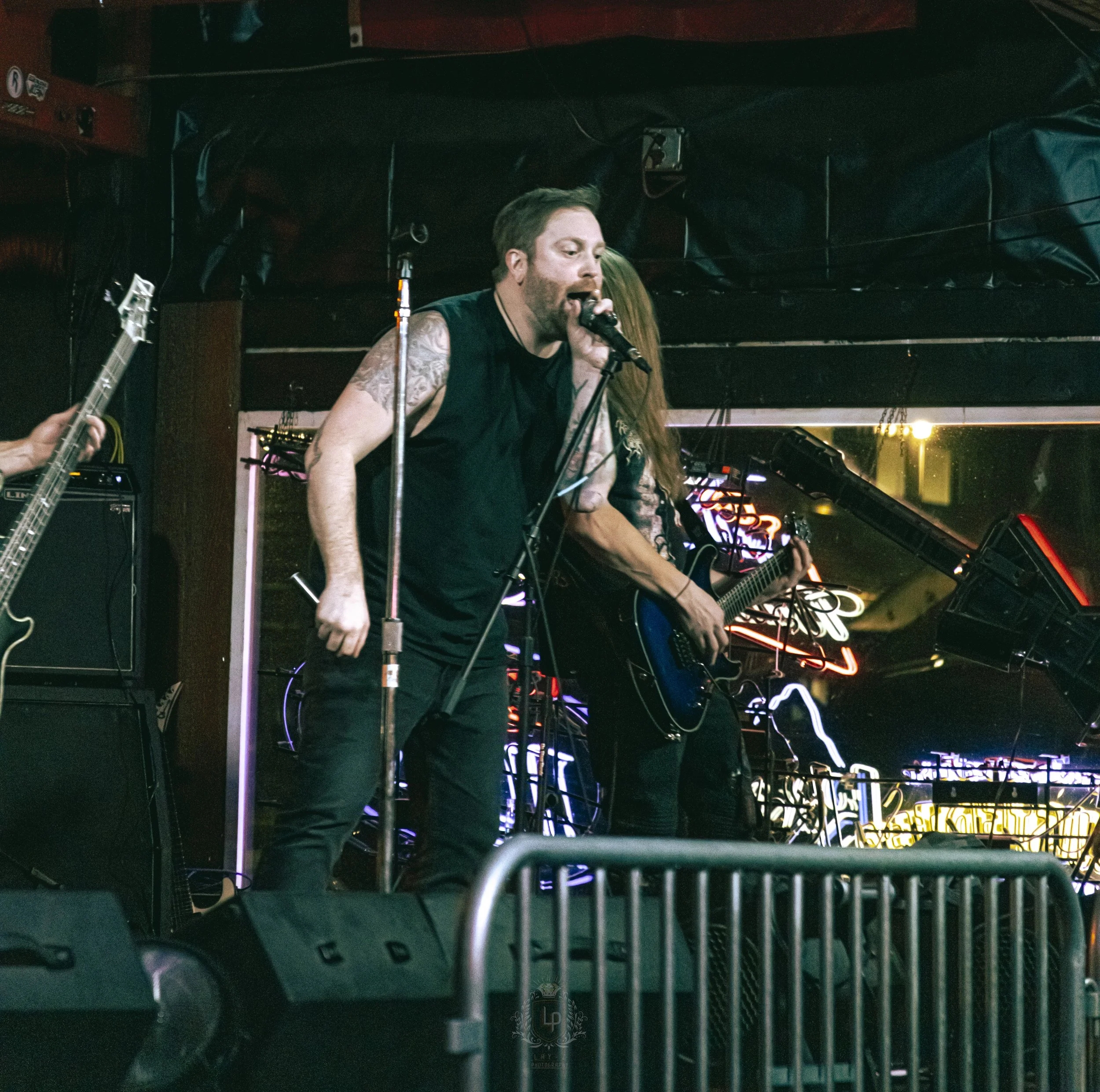 Male vocalist singing into a microphone during a live rock performance at a club with neon signs in the background.