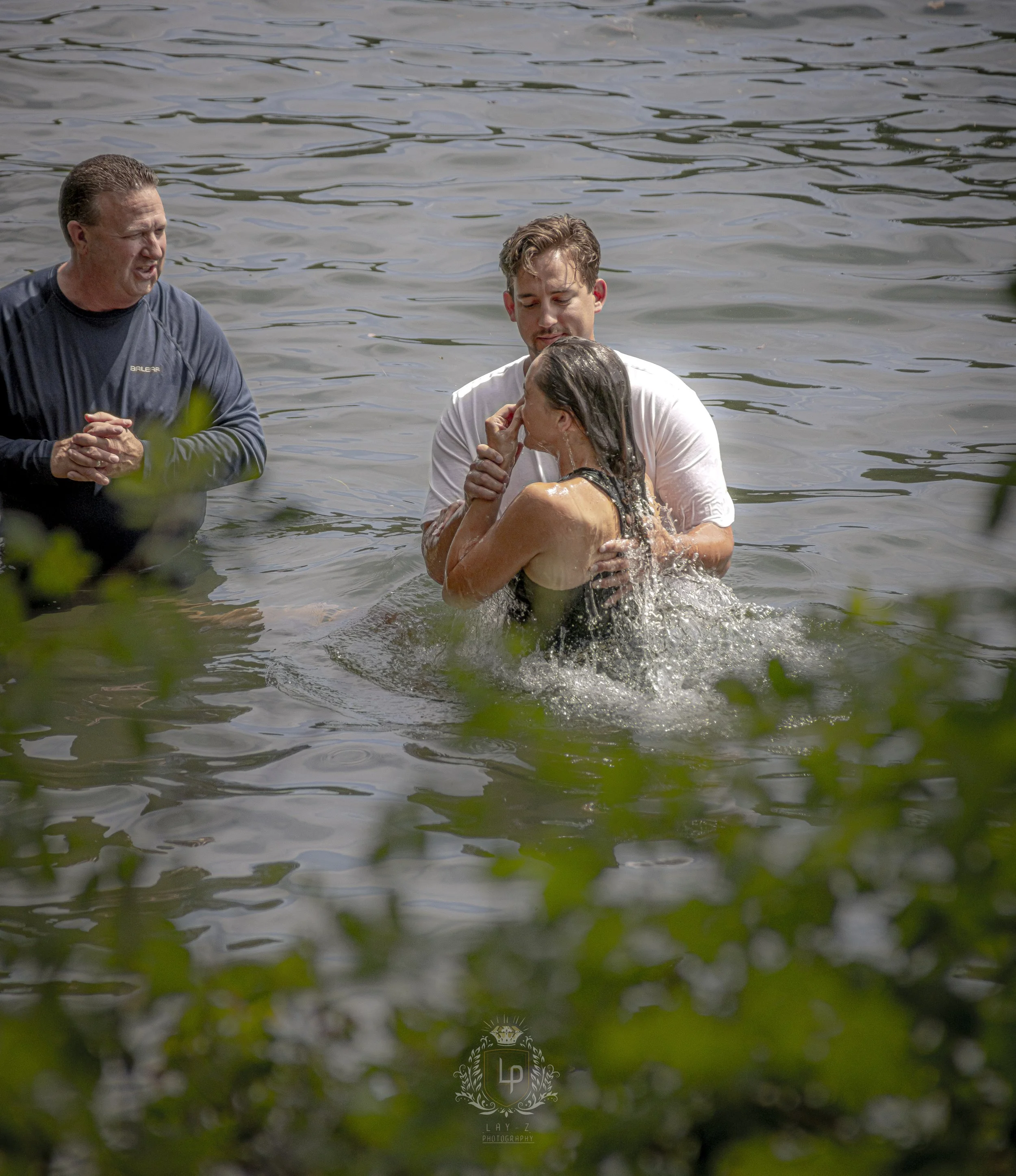 A woman being baptized in a lake by a man, with a priest standing nearby, during daytime.
