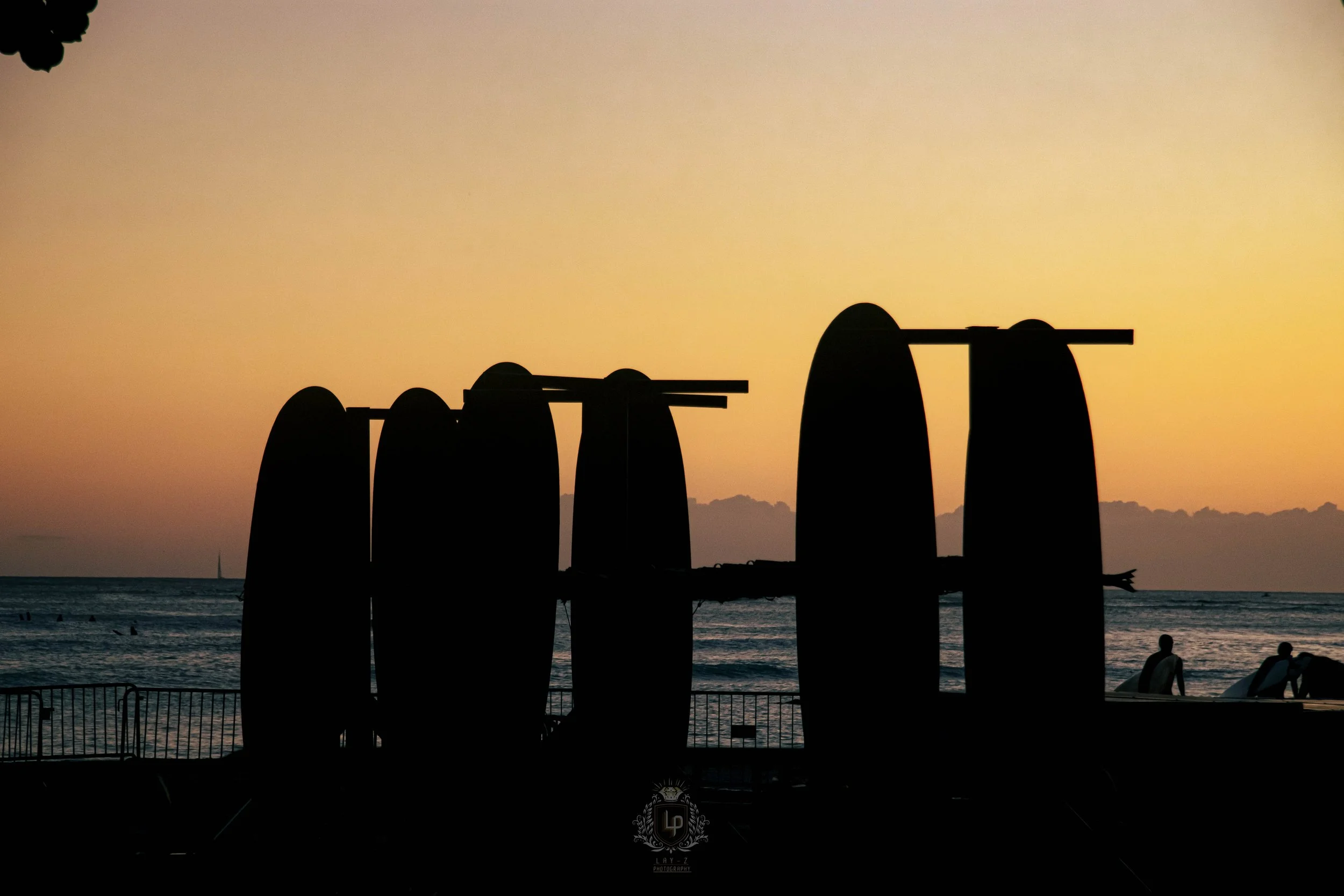 Silhouette of surfboards standing upright on the beach during sunset, with the ocean and people visible in the background.