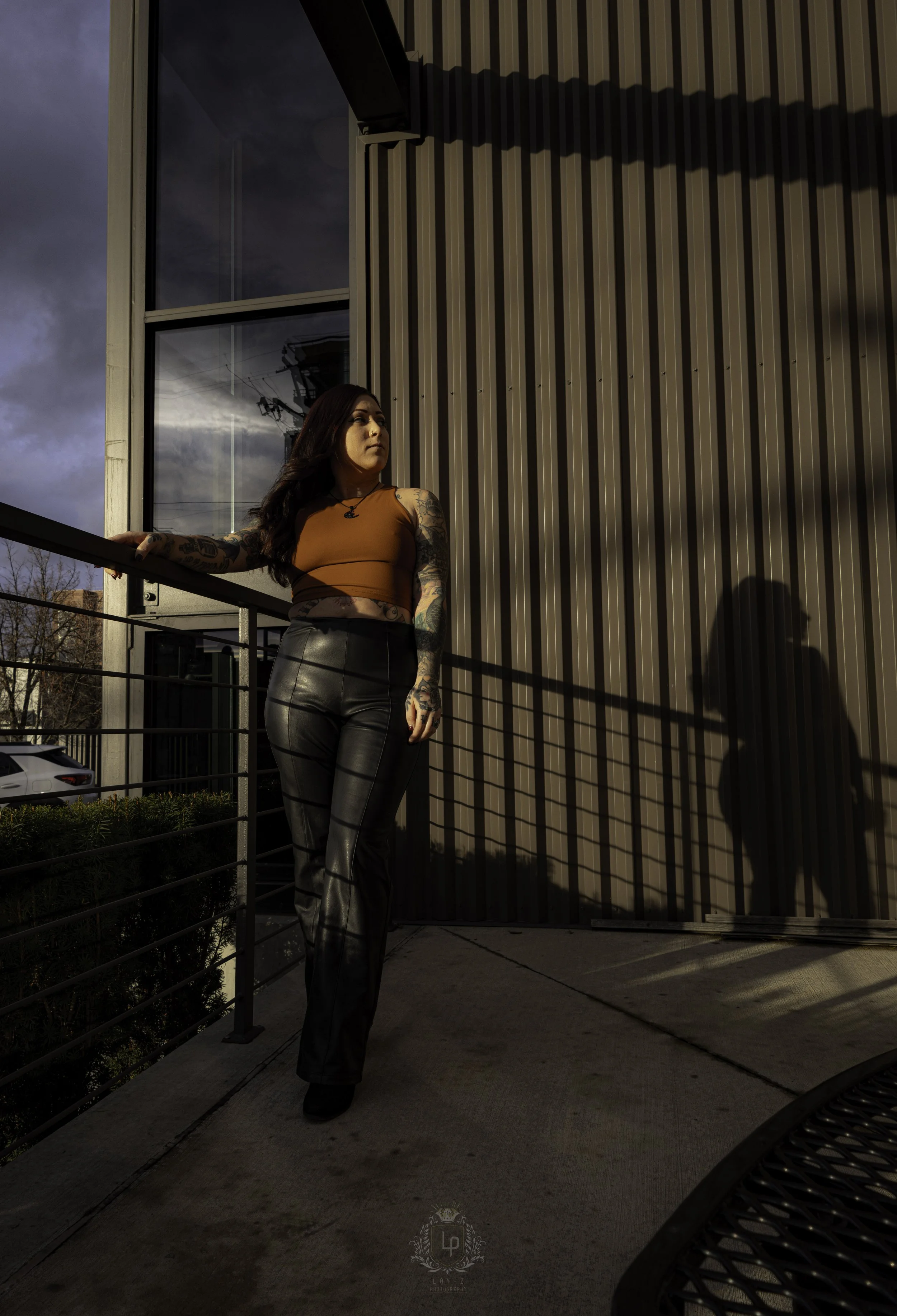 A woman with tattoos wearing a brown crop top and black faux leather pants standing on a balcony, looking to the side with her hand resting on the railing, with dramatic sunlight casting shadows on the wall behind her.