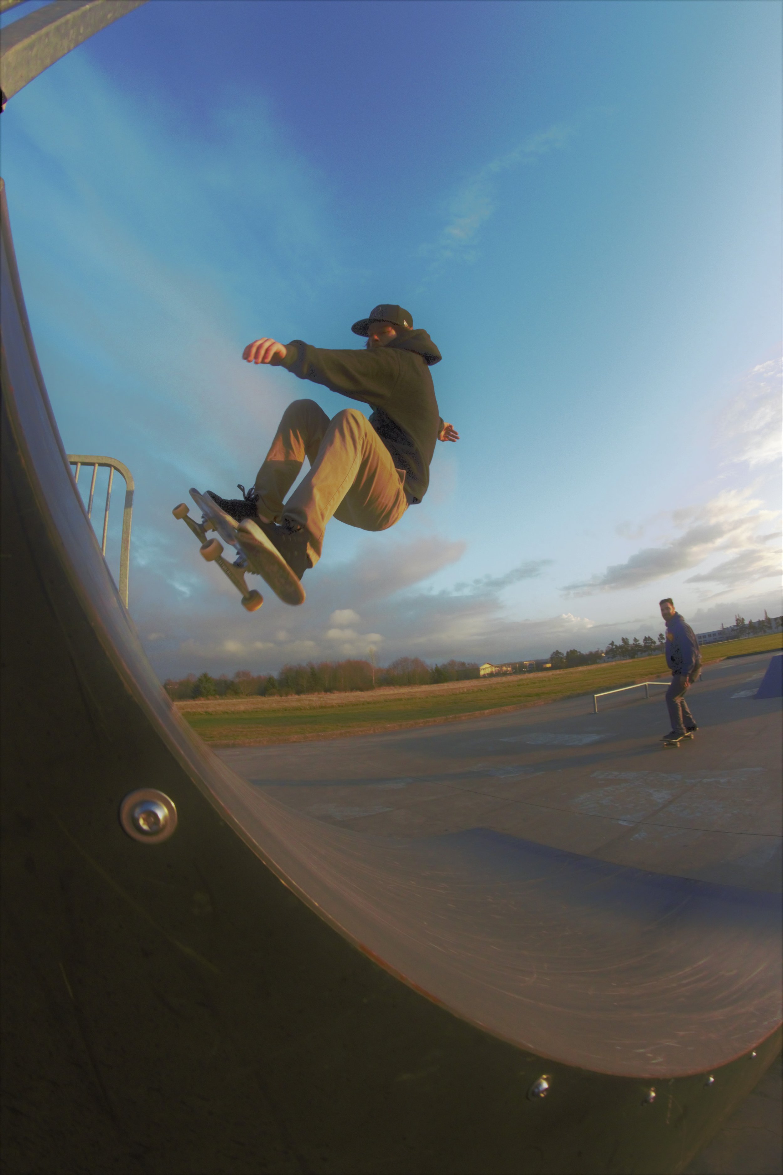A skateboarder in a black hoodie and tan pants performing a trick in the air at a skate park during sunset, with another person skating in the background and an open field beyond.