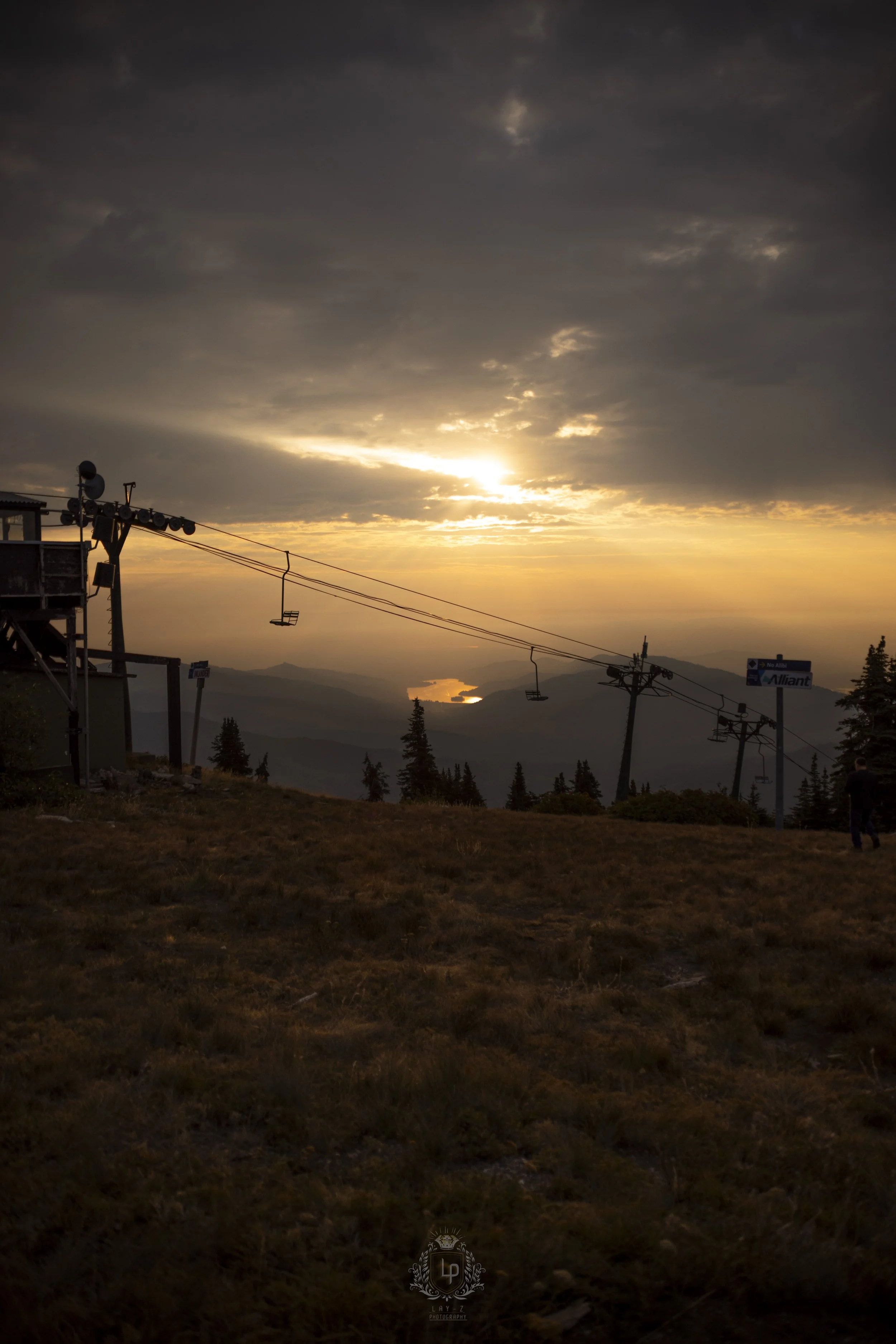 Sunset over mountainous landscape with ski lift cables and chairs in the foreground, and a lake reflecting the sunlight in the distance.