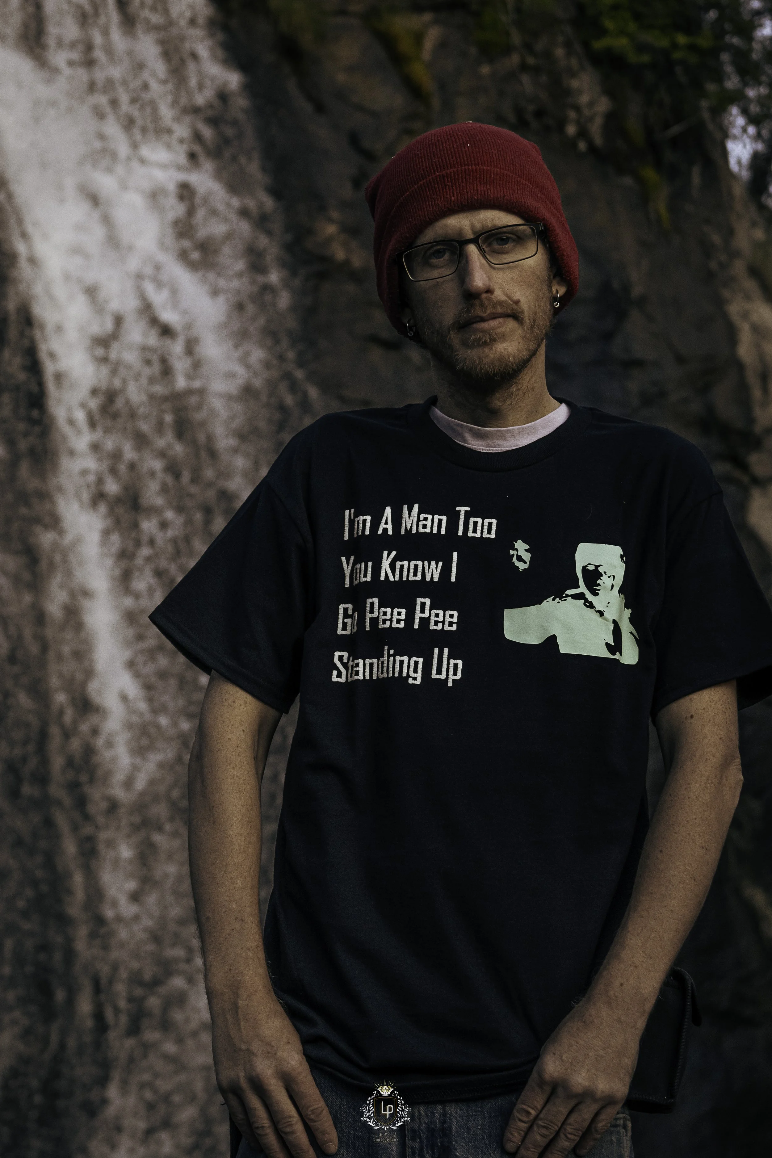 A man wearing a red beanie, glasses, and a black T-shirt with white text and a graphic, standing outdoors near a waterfall.