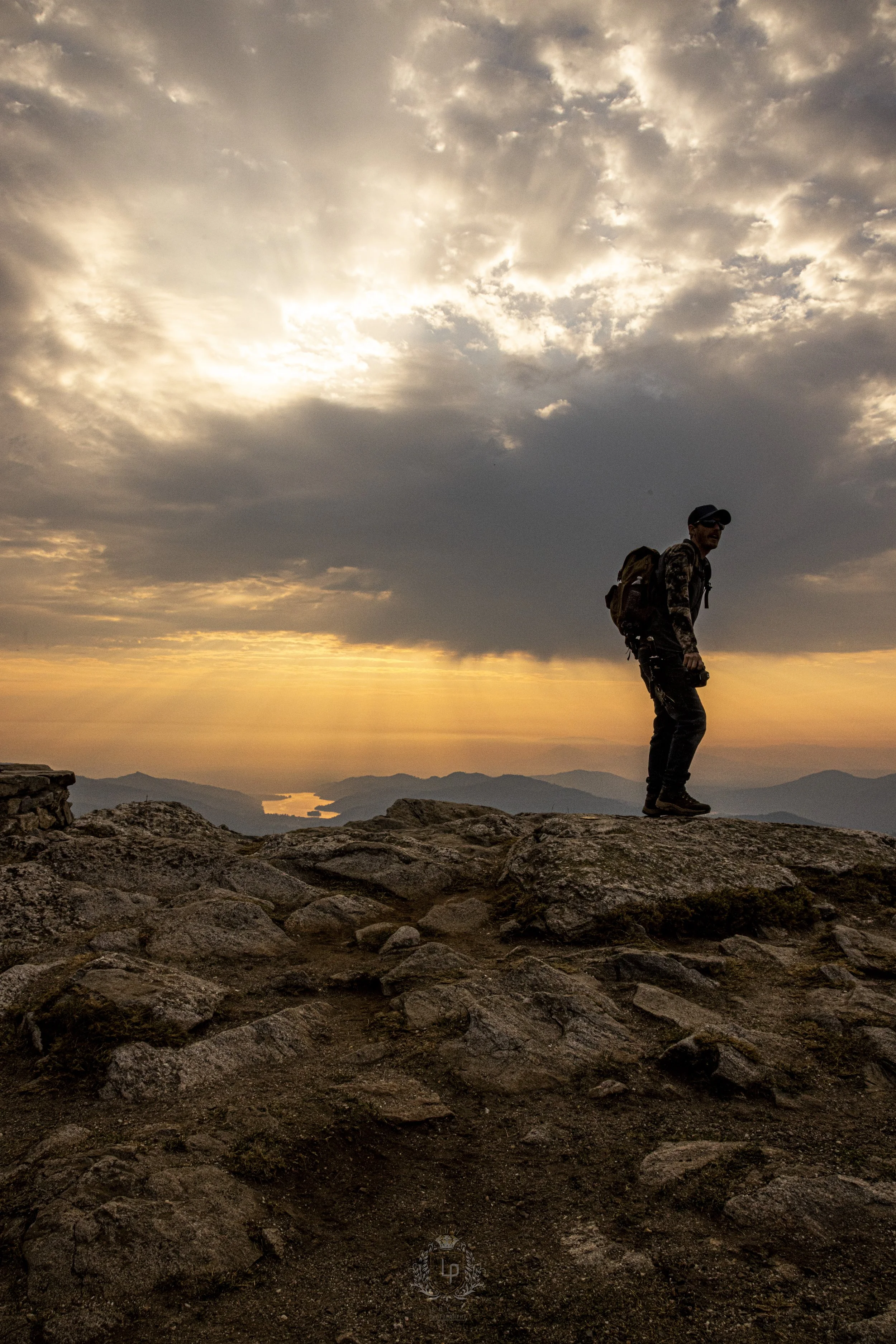 A person standing on rocky terrain during sunset, overlooking mountains and water with dramatic cloudy sky.