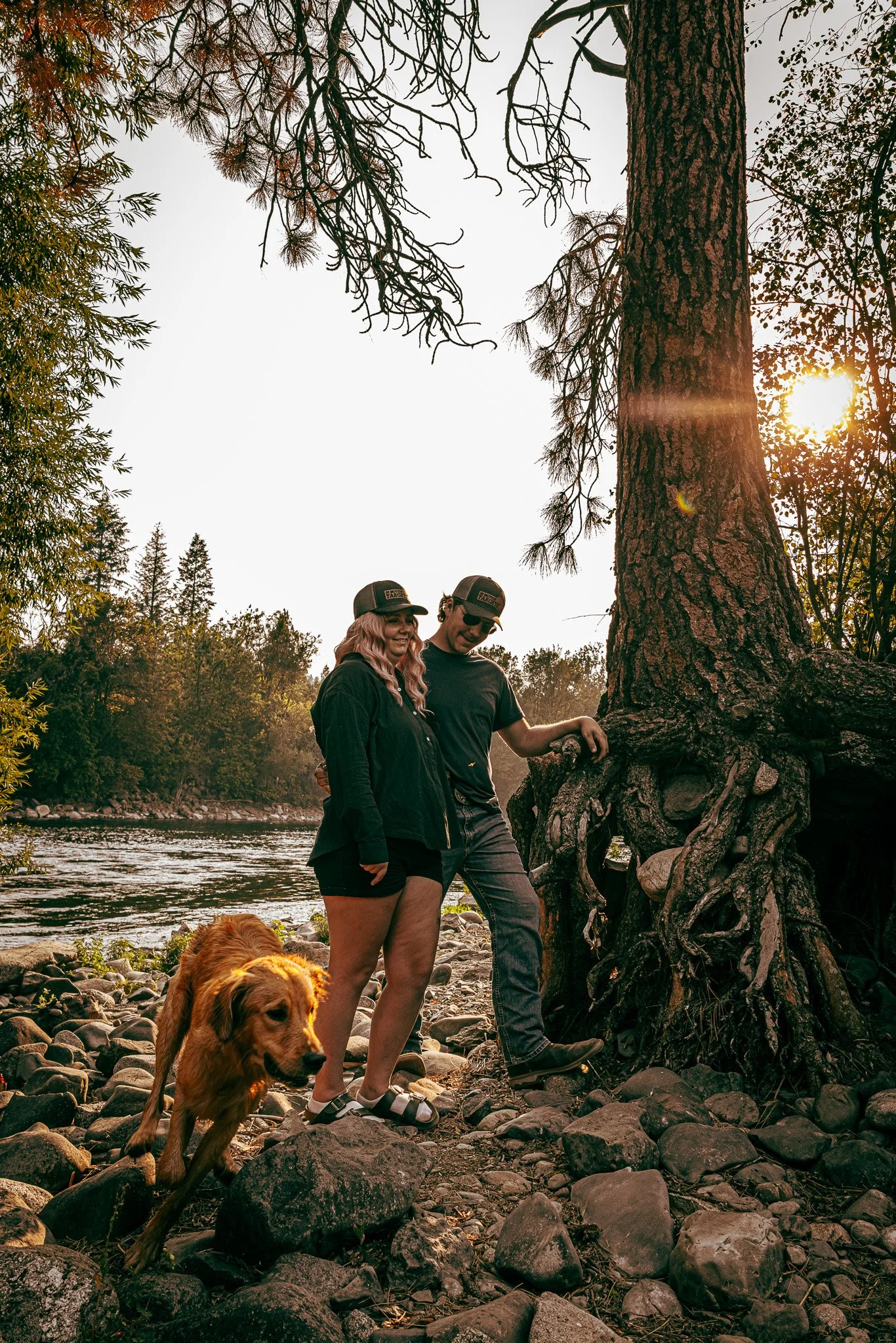 A couple and a dog by a river at sunset, with a large tree and lush greenery in the background.